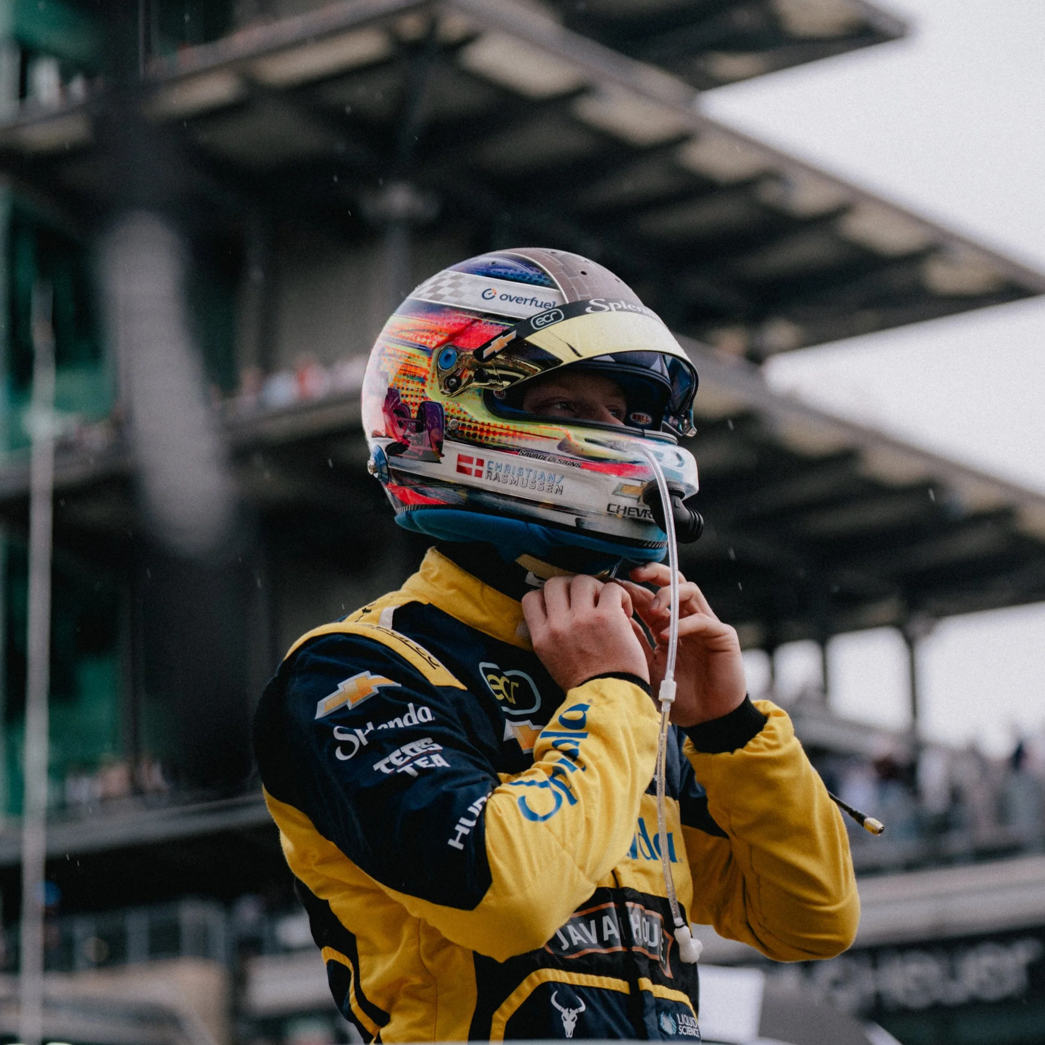 Race car driver Christian Rasmussen in yellow and blue Ed Carpenter Racing racing suit putting on a racing helmet at the Indianapolis Motor Speedway race track.