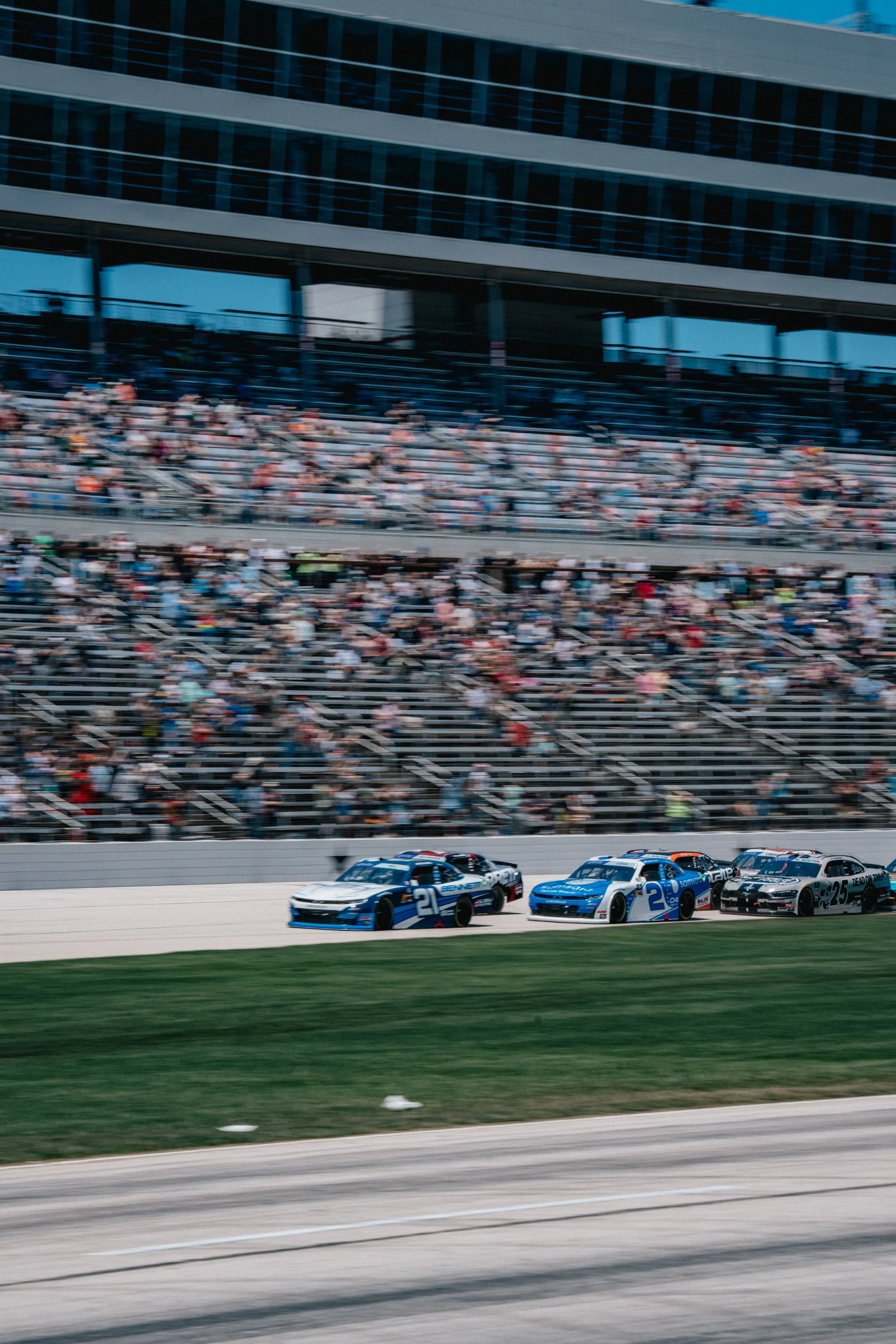 A NASCAR race with three race cars on the track, racing past a large audience in the stands inside a modern stadium during the day.