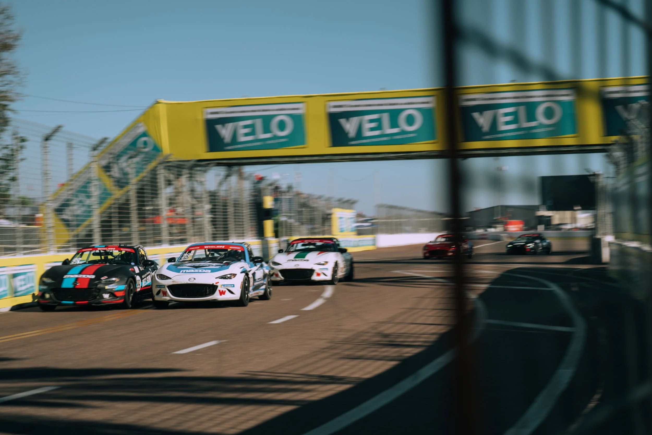 Race cars on a race track, passing under a yellow overhead sign with the text 'VELO', with a fence and stands in the background.