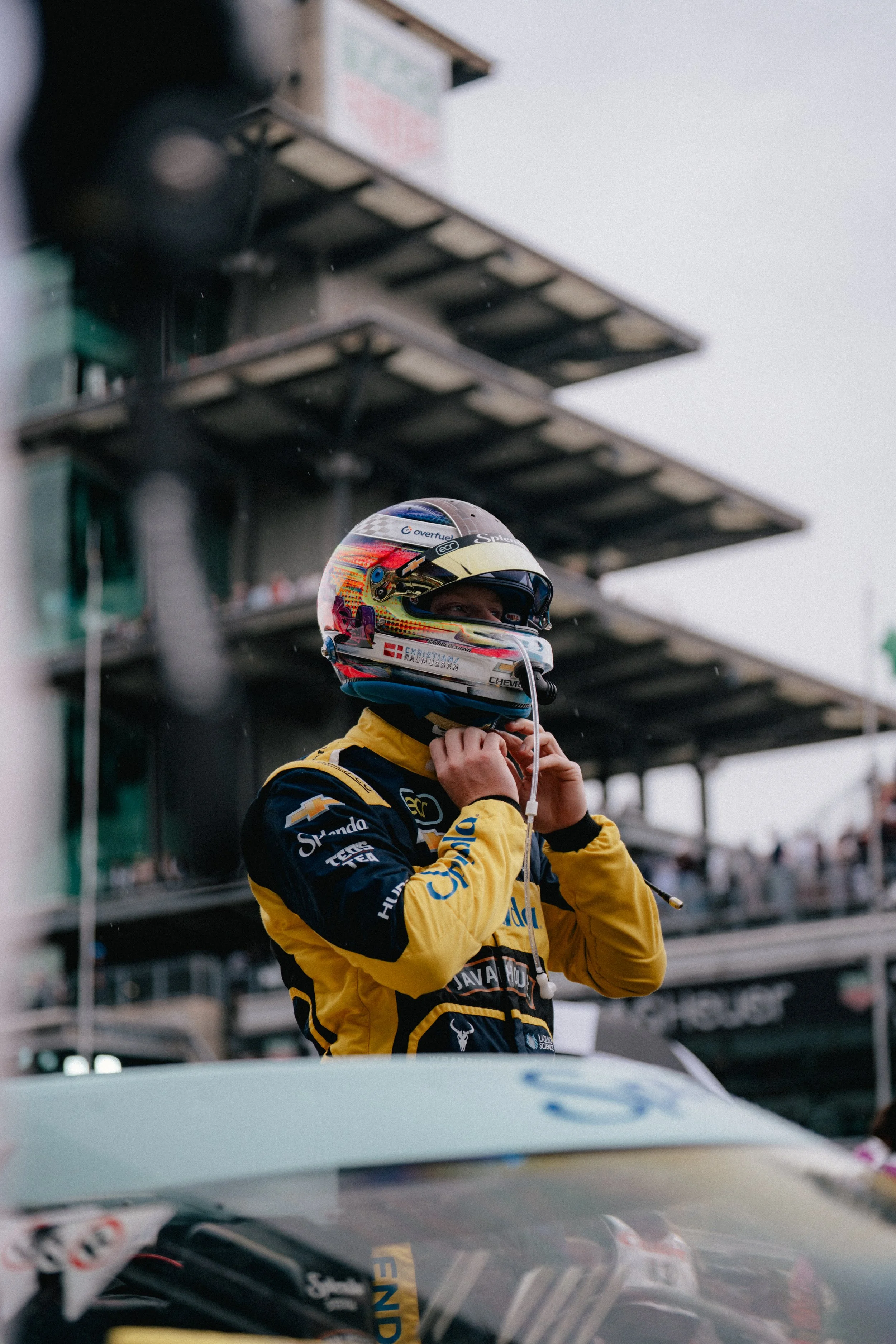 A racing driver wearing a yellow and black racing suit and a colorful helmet is adjusting their helmet strap. They are standing next to a race car with the driver's side visible, and a background of multi-level grandstands at a racetrack.