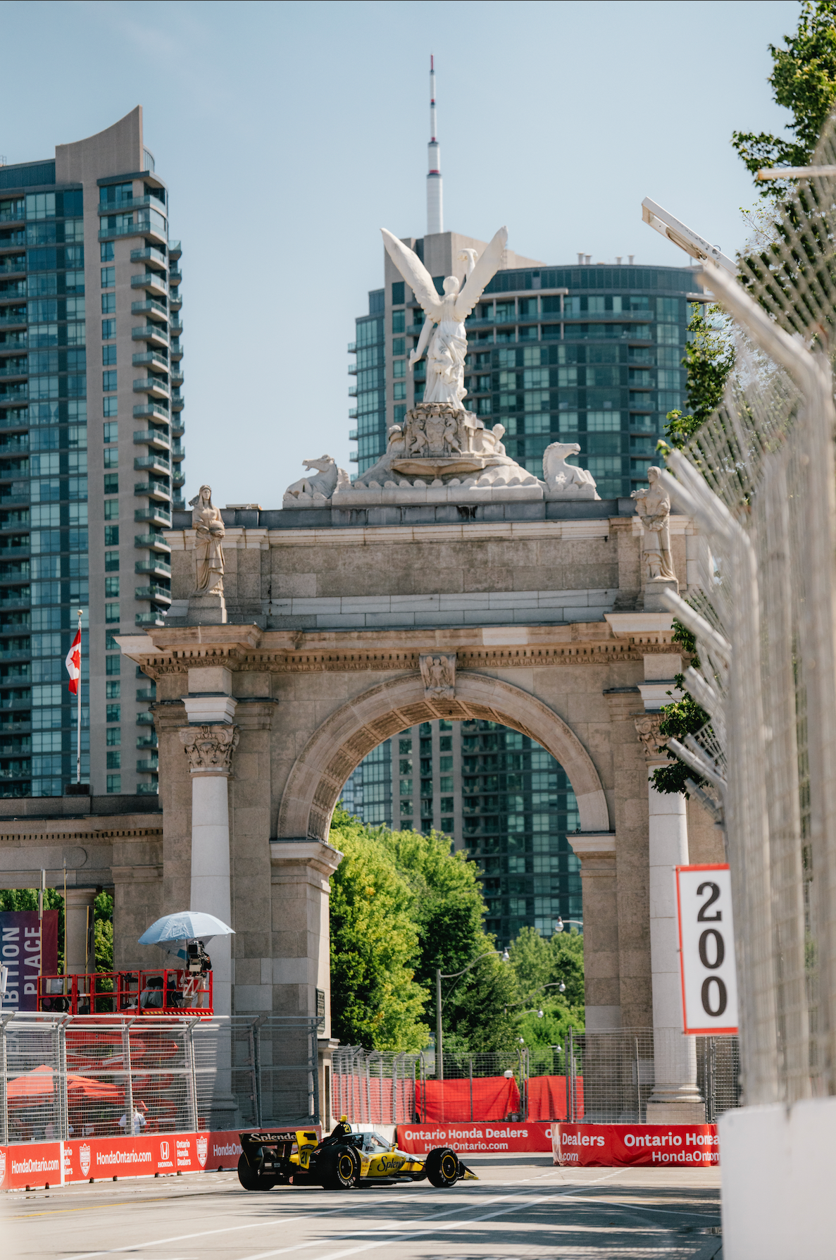 A race car on a track passing beneath a historic stone archway with statues and a winged figure on top, surrounded by modern high-rise buildings and a blue sky.