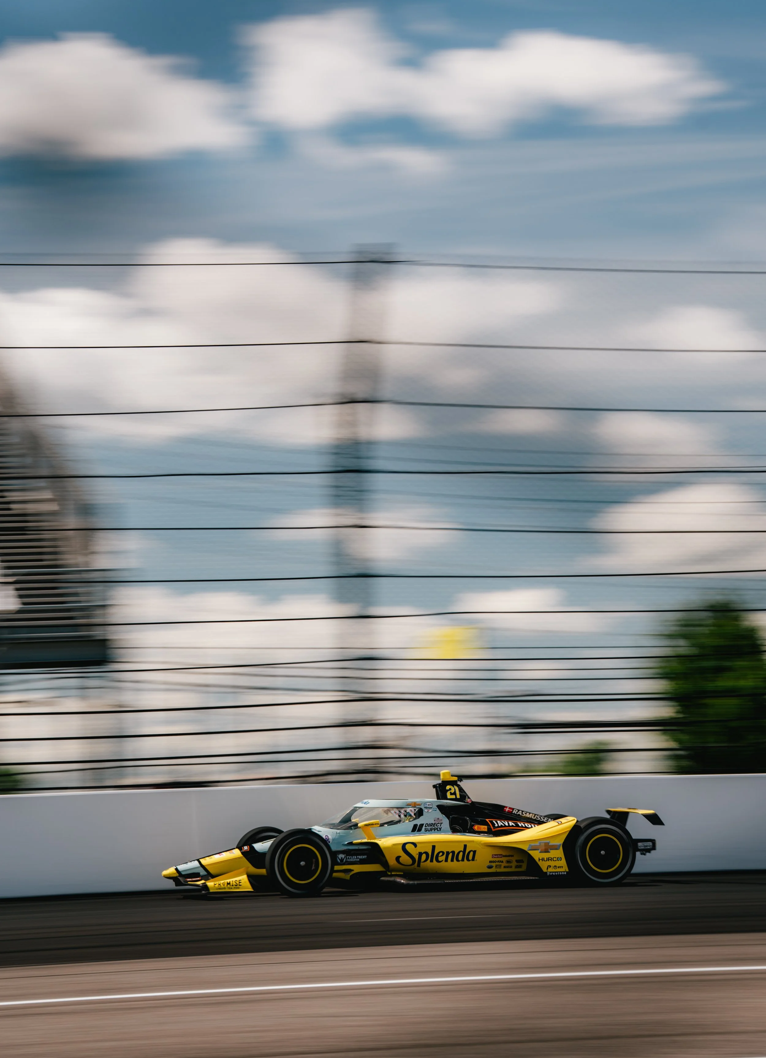 A yellow and black IndyCar race car speeding on a track with a blurred background of a fence and sky.