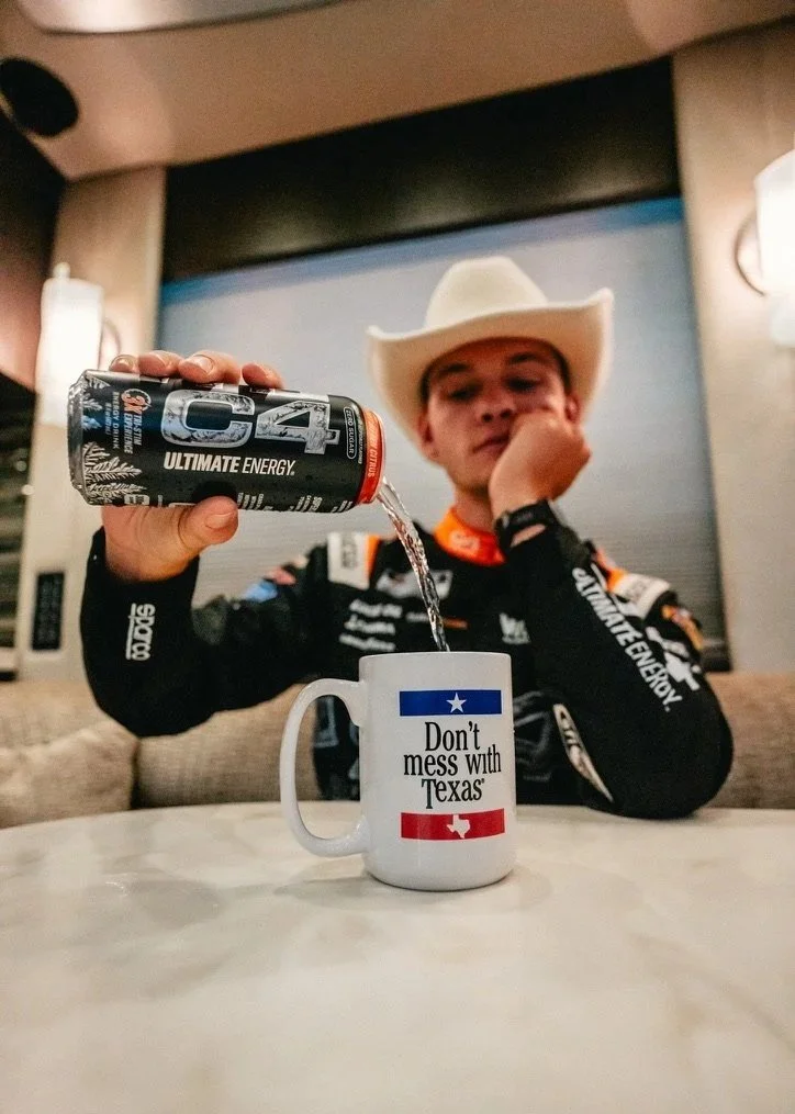A person wearing a cowboy hat and racing suit pouring an energy drink into a coffee mug with Texas-themed slogan.