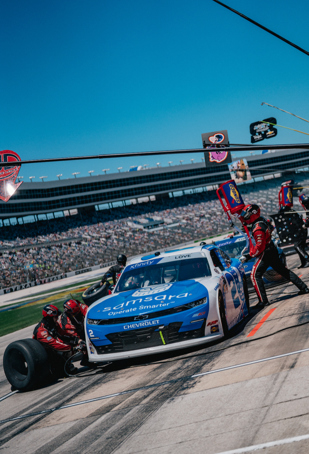 Race car in the pit stop with crew members changing tires during a NASCAR race at a large outdoor stadium.