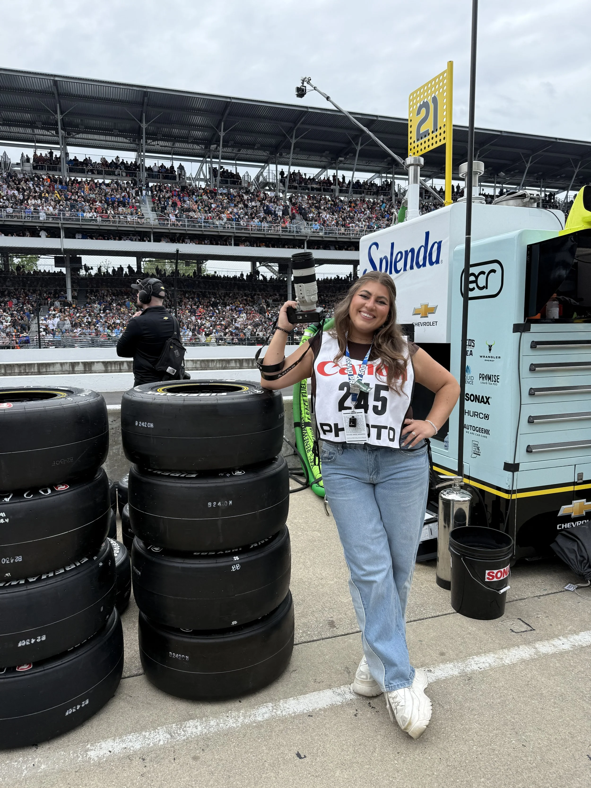 A smiling woman standing at a racing event, holding a camera with a large lens. She's wearing a white bib with the number 245, light blue jeans, and white sneakers. Behind her are stacks of tires, a pit box labeled 'Splenda' and a large crowd in the grandstands.