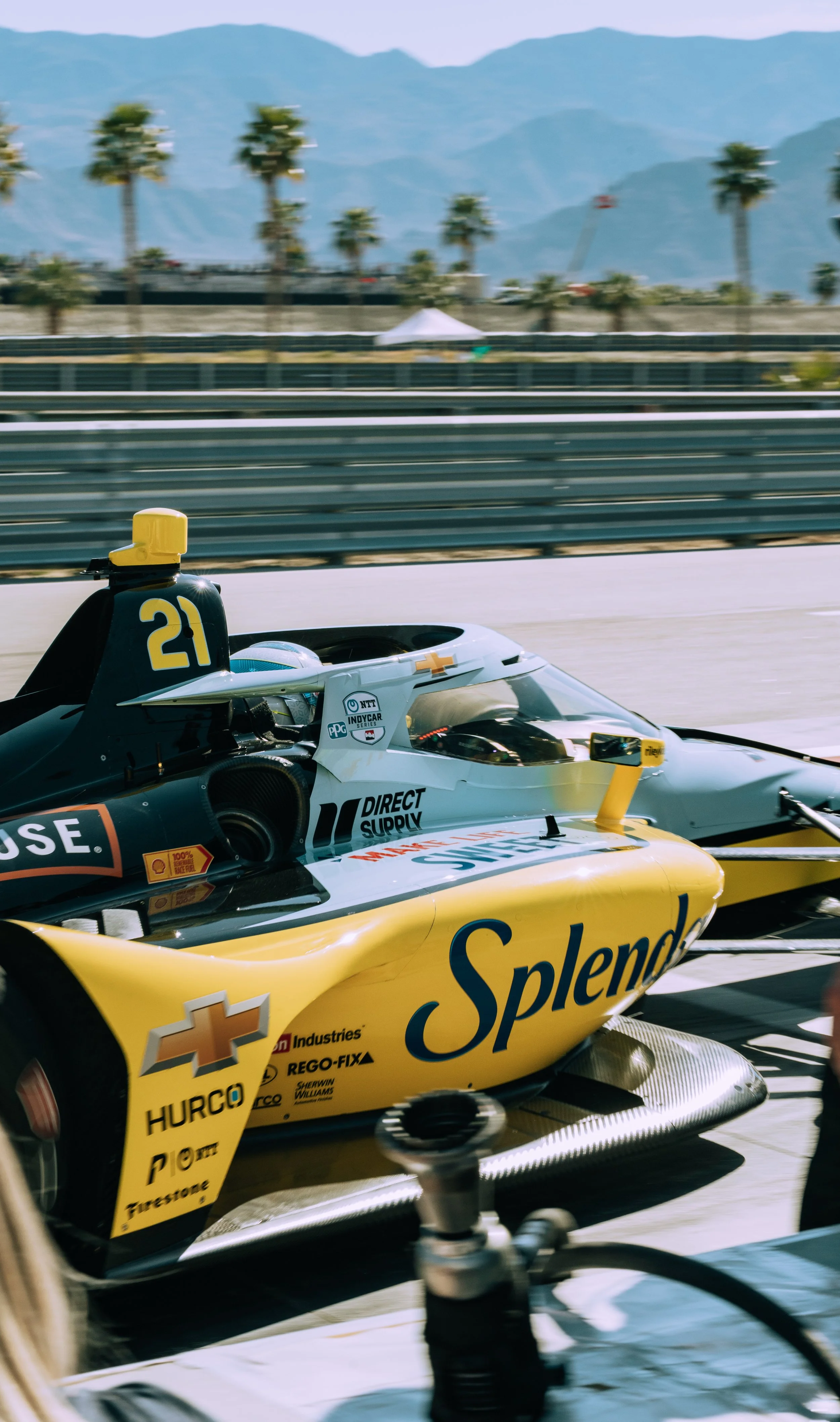IndyCar race car on the track with sponsor logos, a yellow and black color scheme, and a mountainous landscape in the background.