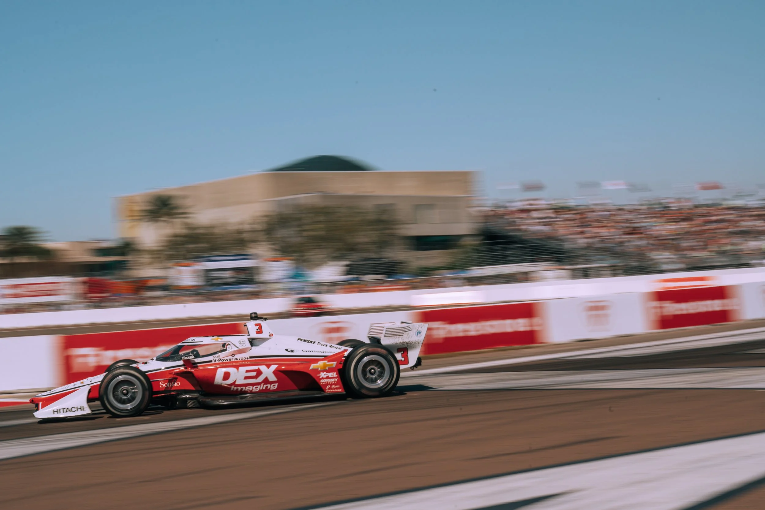 A race car speeding on a race track during a race with a grandstand filled with spectators in the background.