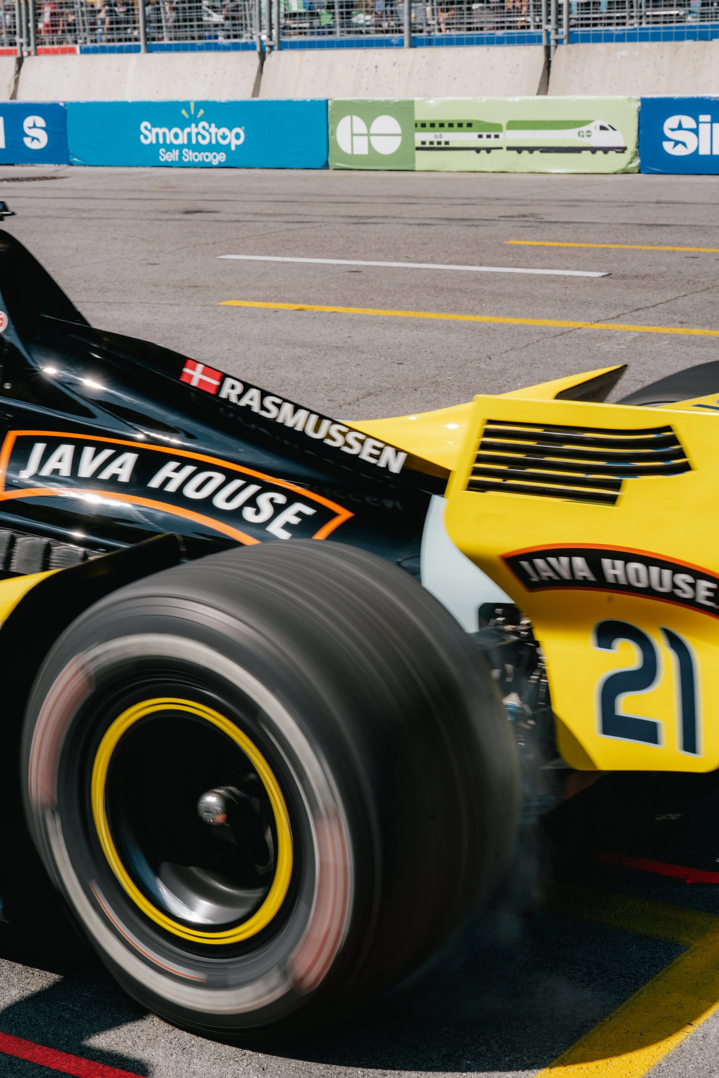 Close-up of a race car tire with smoke, yellow and black body with sponsor decals including Java House and RASMUSSEN, number 21, on a race track with barriers and advertisements in the background.