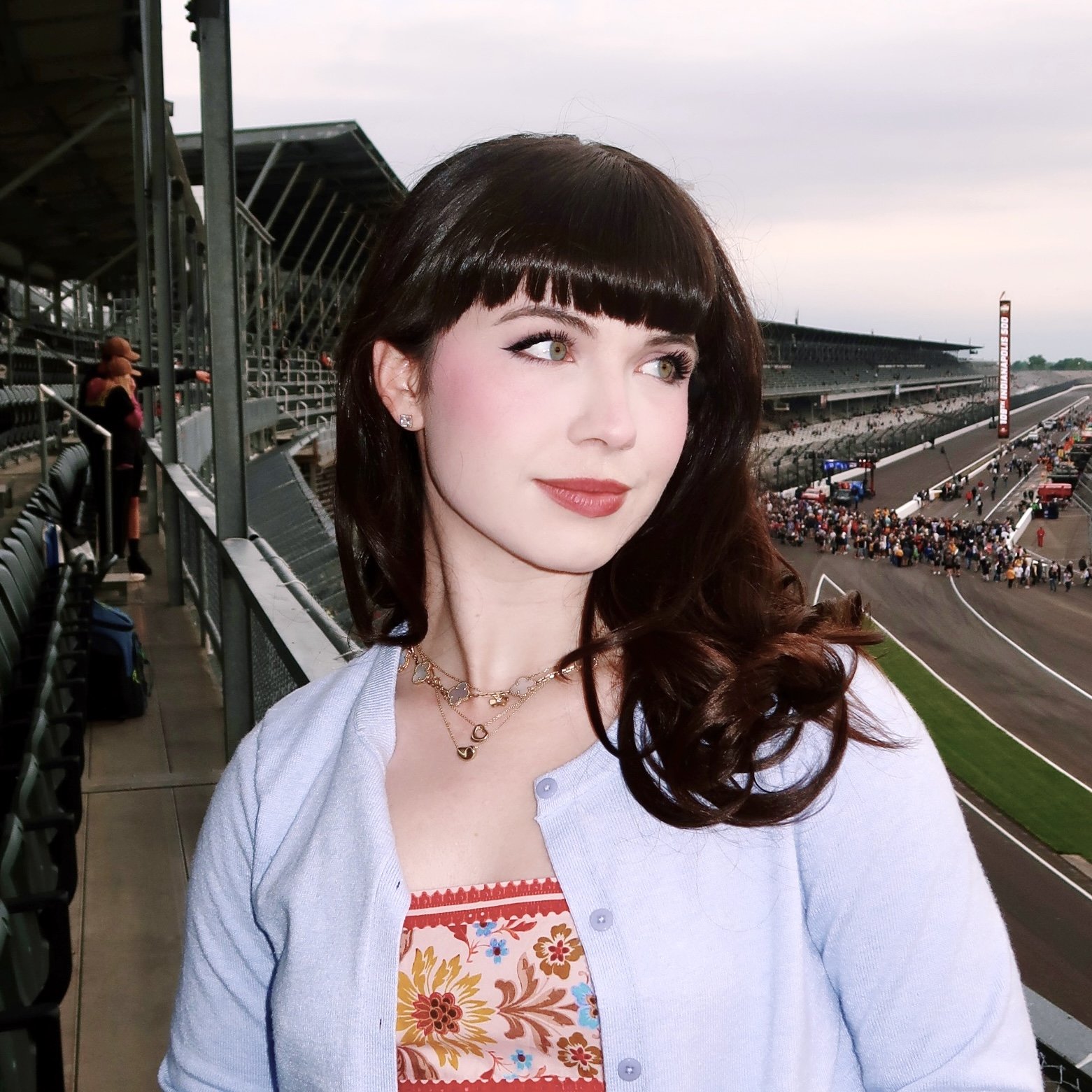 A young woman with dark brown hair and bangs, wearing a light blue cardigan and layered necklaces, posing at a race track with a crowd and cars in the background.