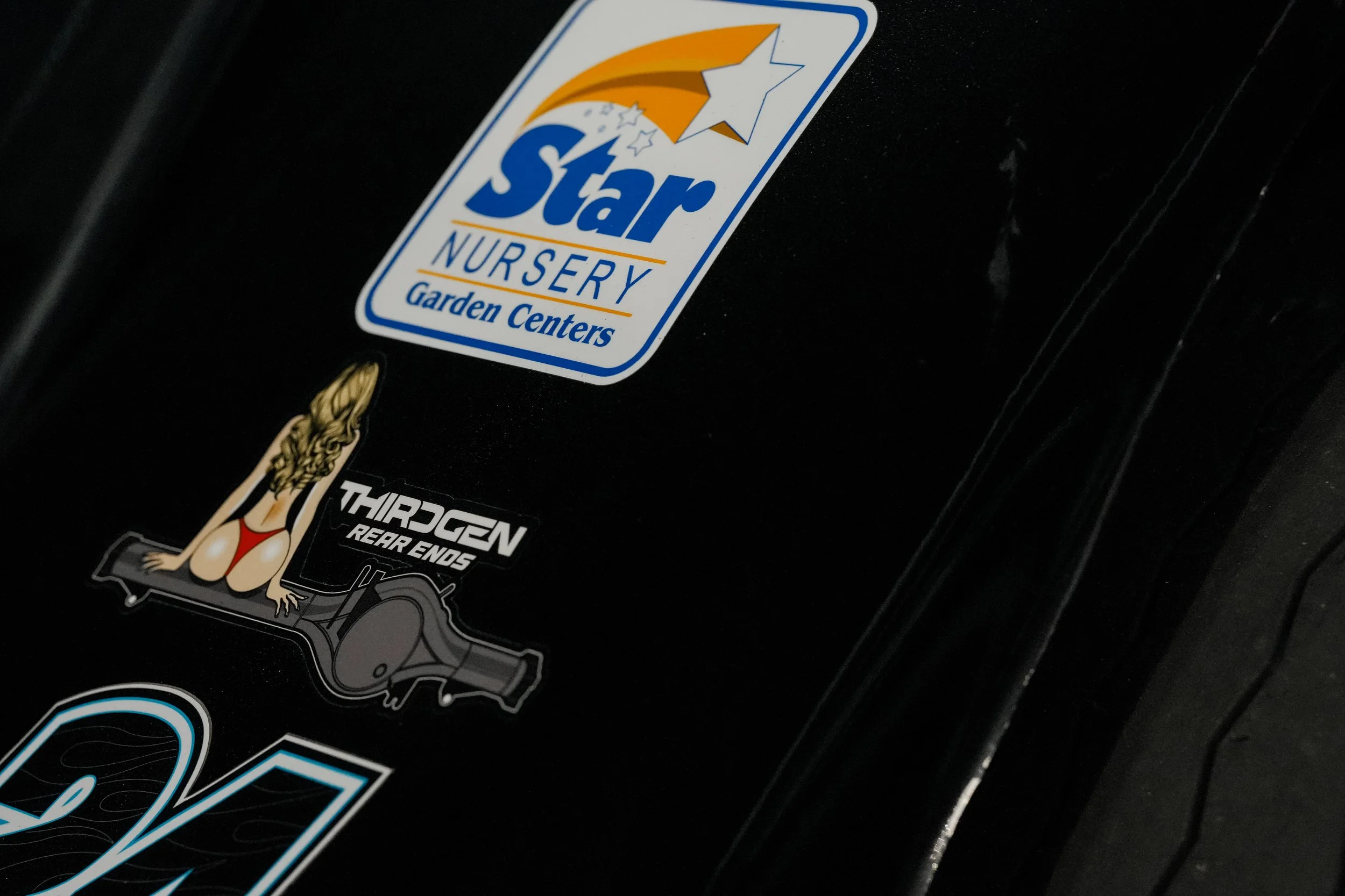 Close-up of car stickers on a black surface, including a Star Nursery Garden Centers logo with a star and shooting star design, and a sticker of a woman in a red bikini with long blonde hair sitting next to a turbo engine with rear end label.