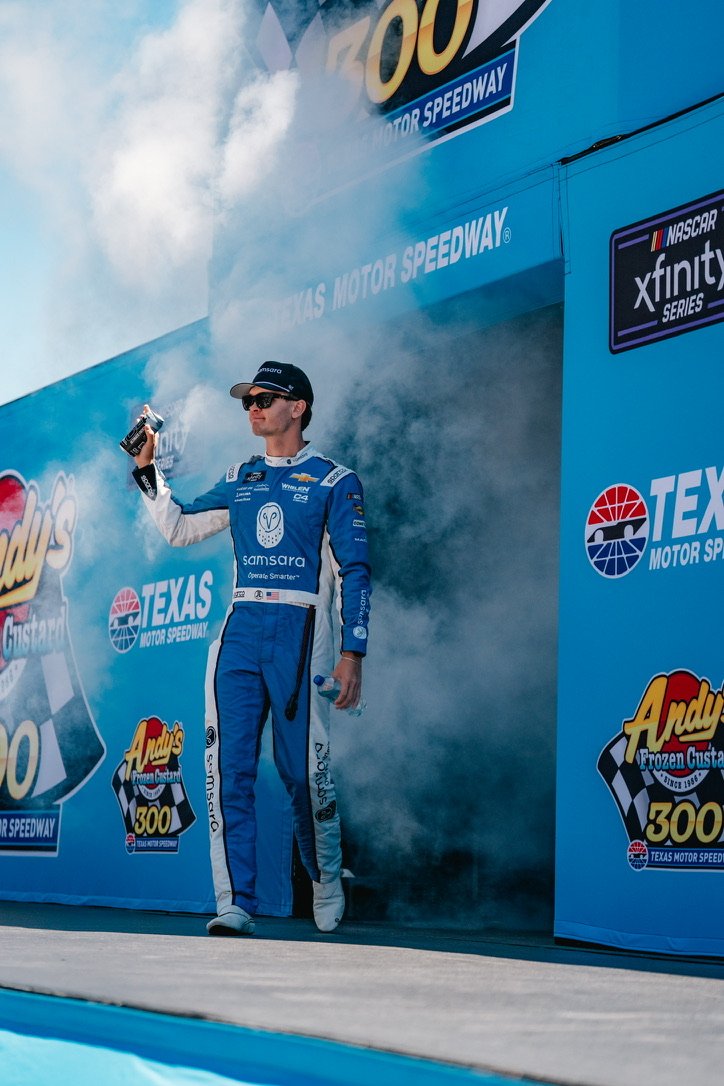 Race car driver in blue racing suit standing on podium at Texas Motor Speedway, holding a bottle and spraying champagne with smoke in the background.