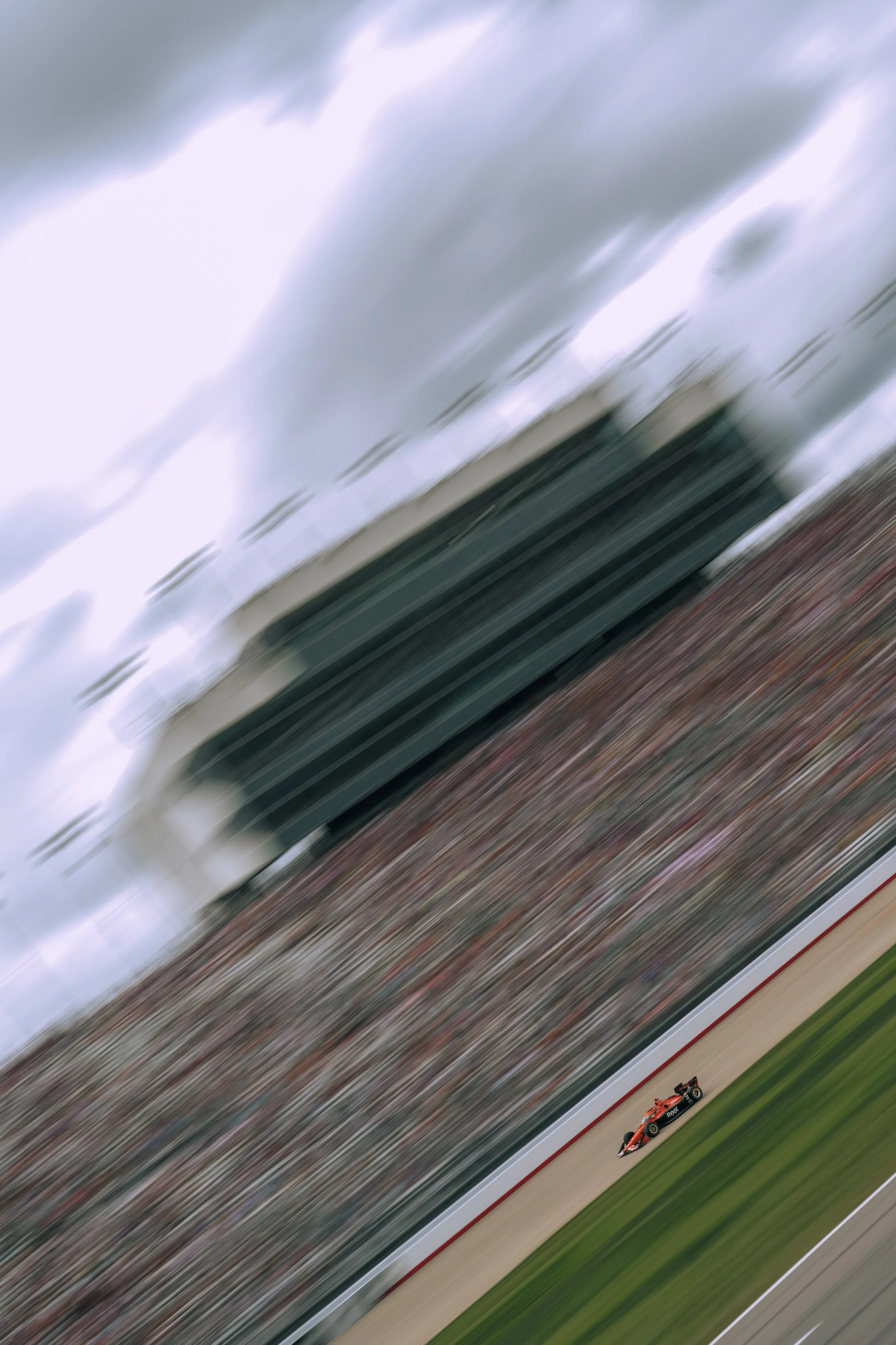 A racing car speeding on a racetrack with it blurred from motion, and a grandstand full of spectators in the background, with a cloudy sky overhead.