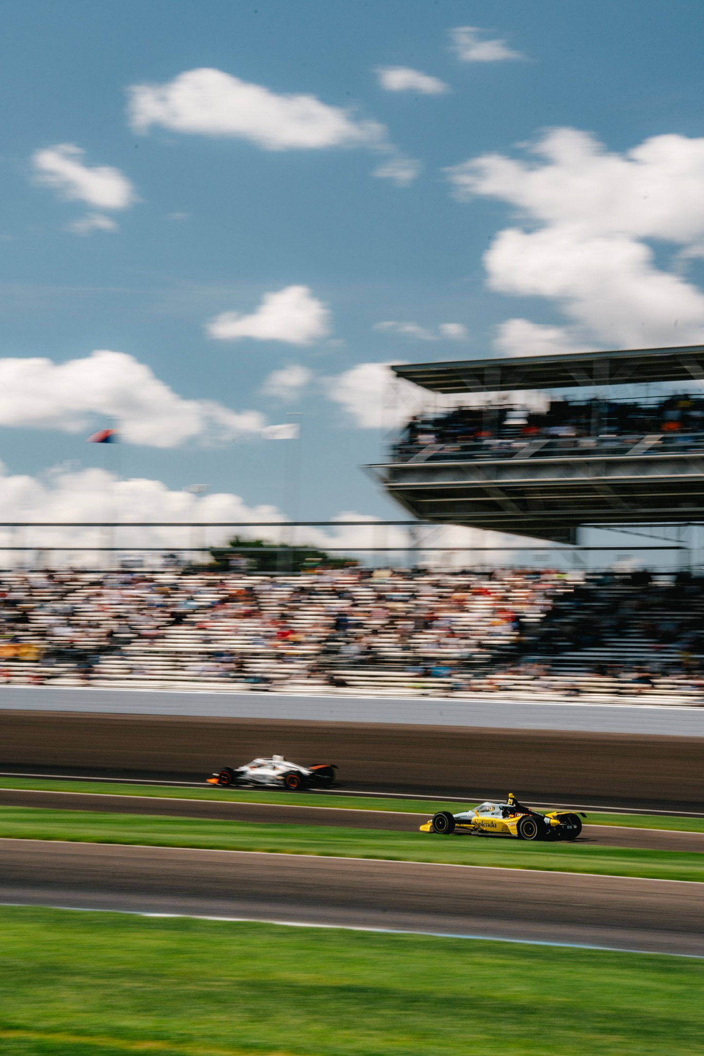 Race cars speeding on a race track with a large crowd in the stands and a partly cloudy sky above.