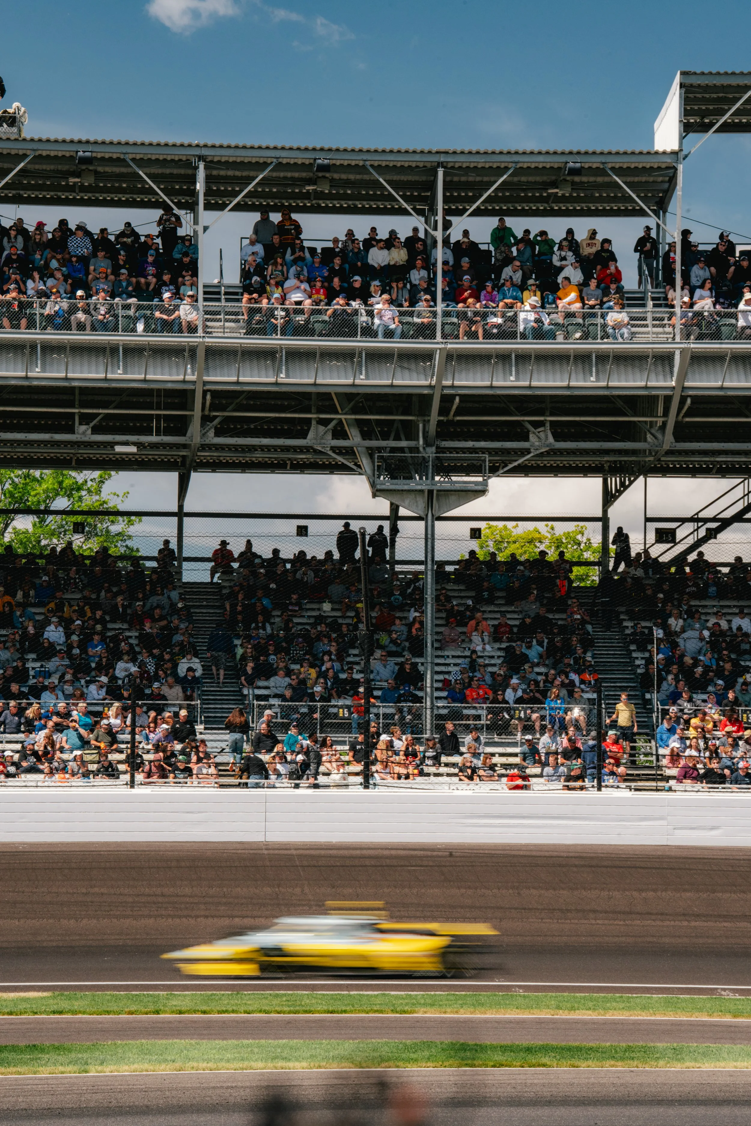 Speeding yellow race car on a dirt track with grandstands filled with spectators in the background.