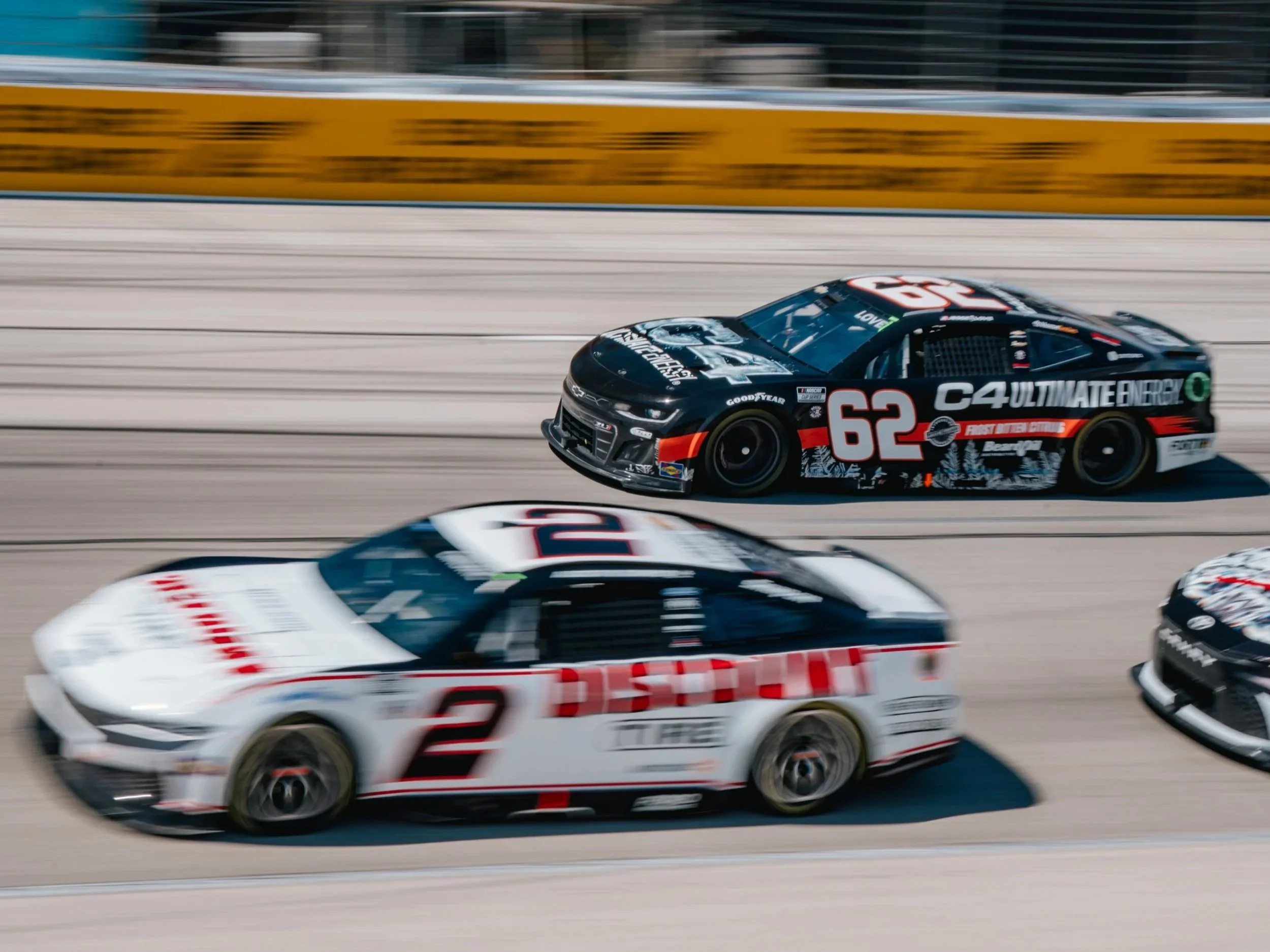 Race cars on a racetrack, including a black and red car numbered 62 and a white and black car numbered 2, racing closely.