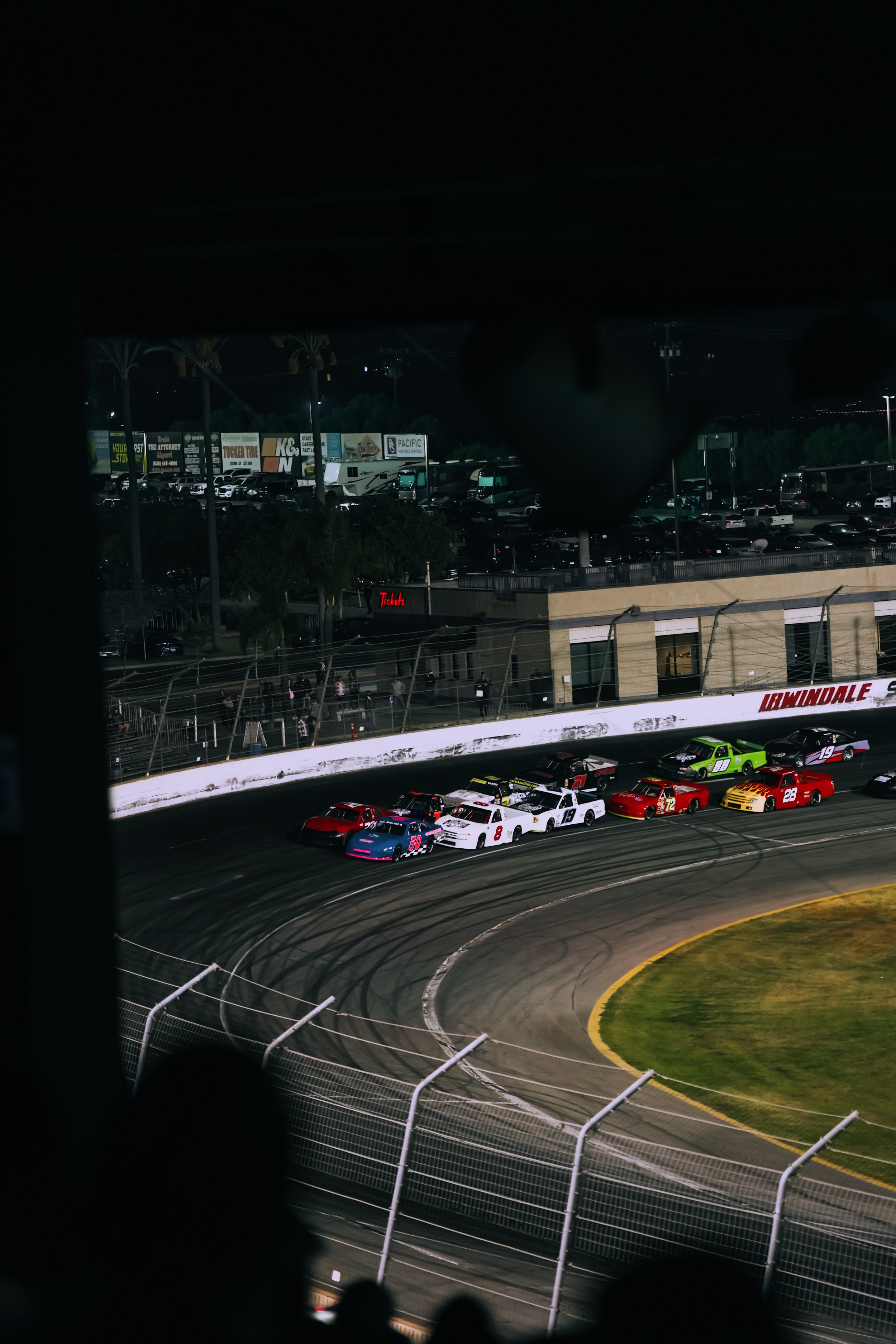 Nighttime race at a racetrack with multiple race cars on the track, surrounded by a fence and grandstands, with parked trucks and billboards in the background.