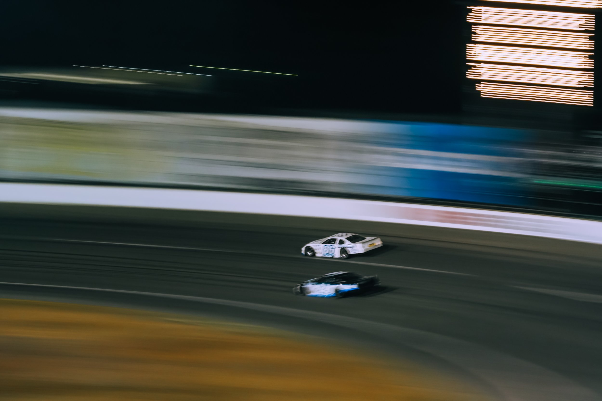 Nighttime NASCAR race with two race cars speeding on the track under illuminated scoreboard, some motion blur.