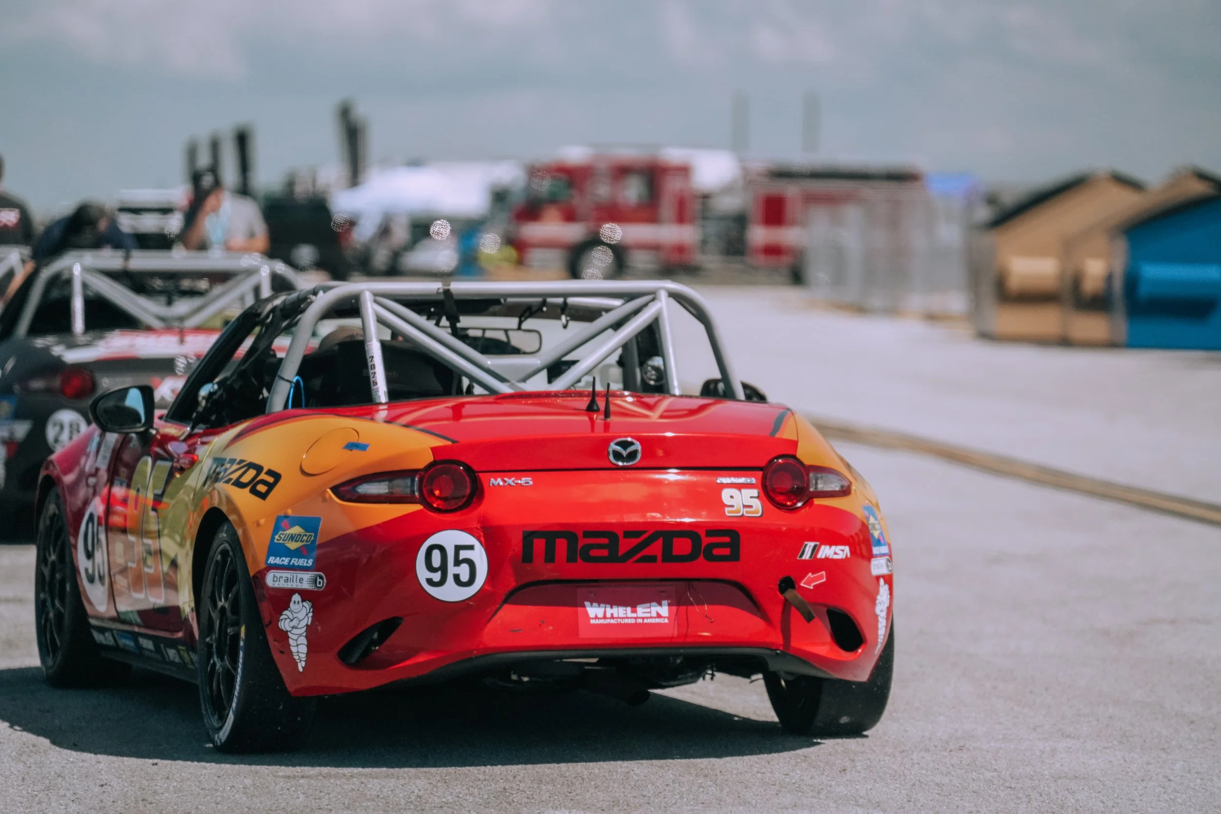 Red Mazda race car with the number 95 on it parked on a race track, with other race cars and crew members in the background.
