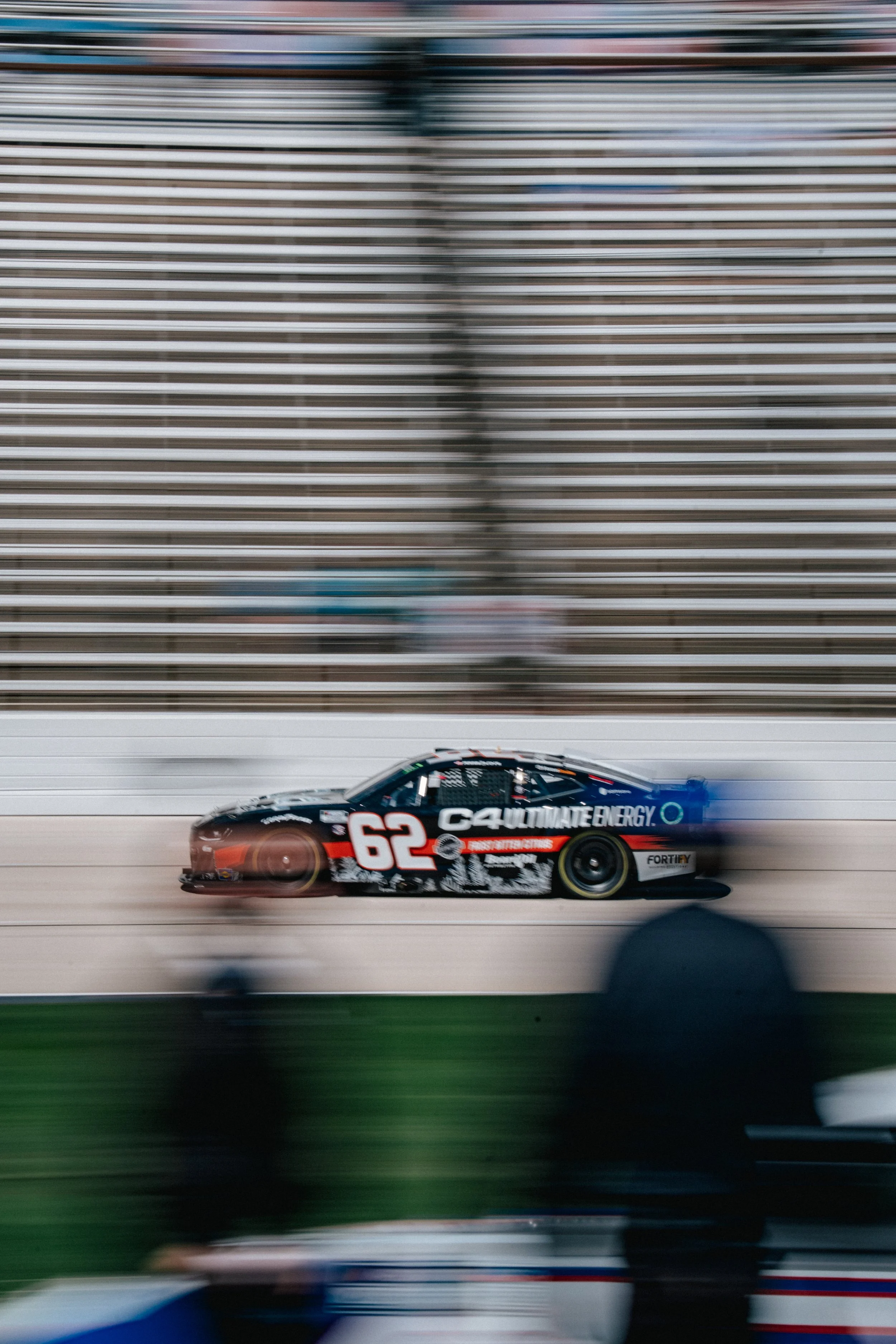 Race car number 62 speeding on the track with motion blur, wall and spectators blurred in the background.