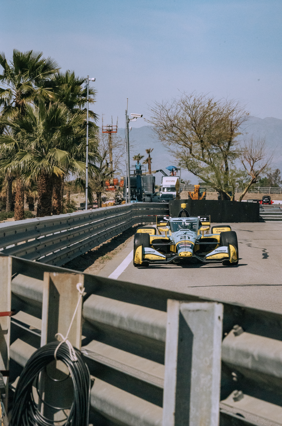 A yellow and black IndyCar race car on the track, next to a safety barrier with some equipment and trees in the background.