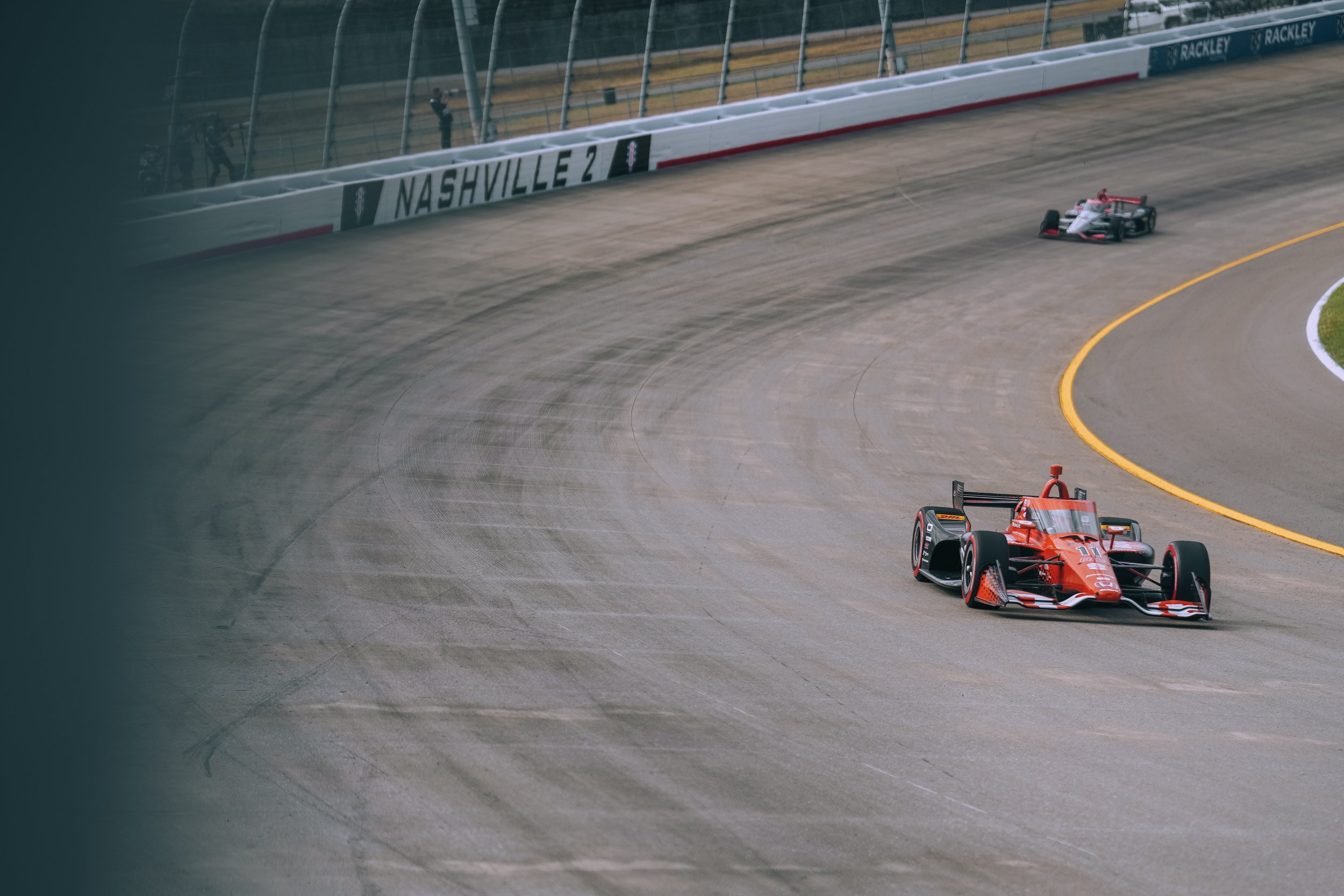 Two open-wheel race cars on a race track with racing barriers and the sign Nashville 2