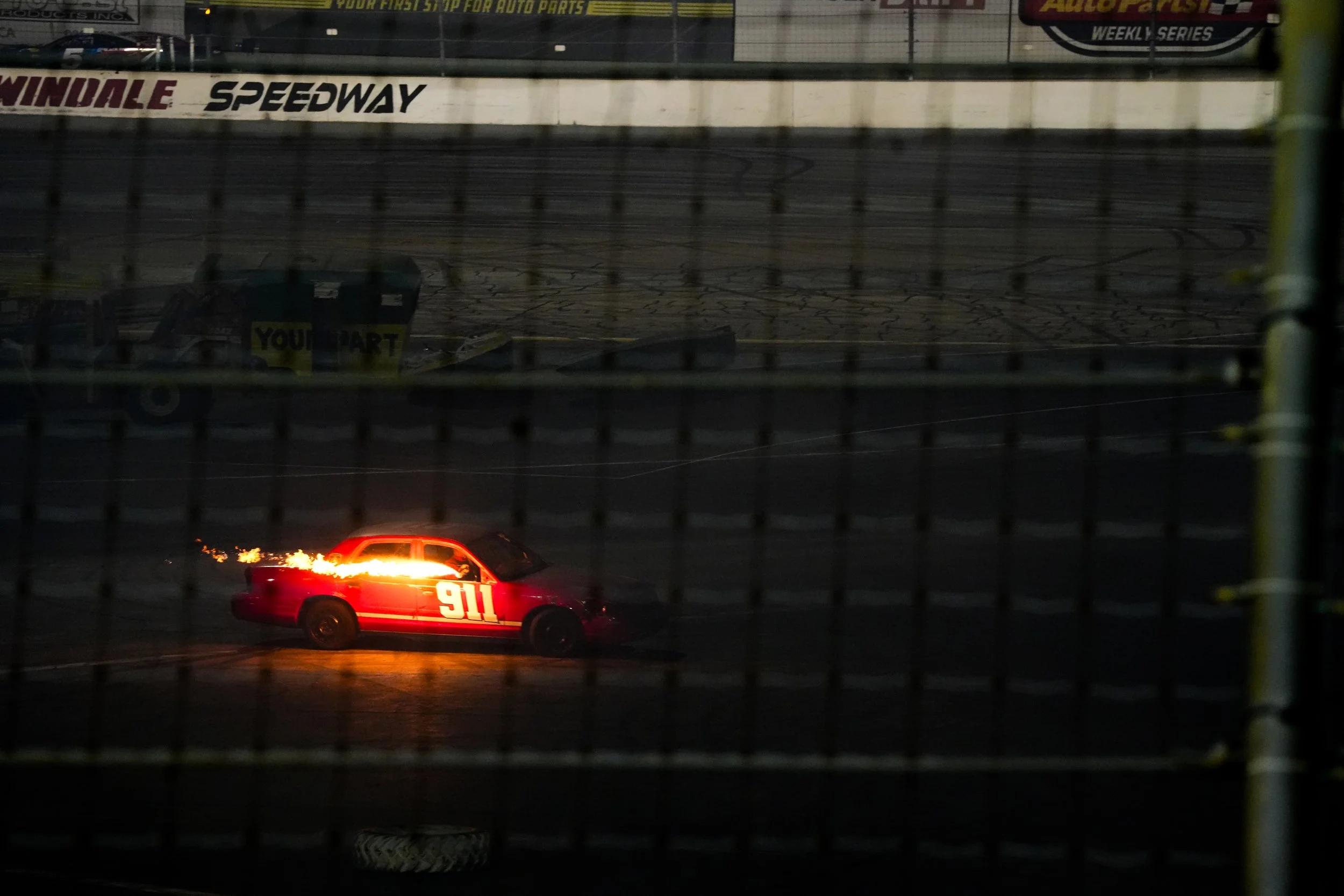 A red night-time racing safety vehicle with the number 911 on its side parked on a racing track, seen through a wire fence with a blurred background of a speedway.