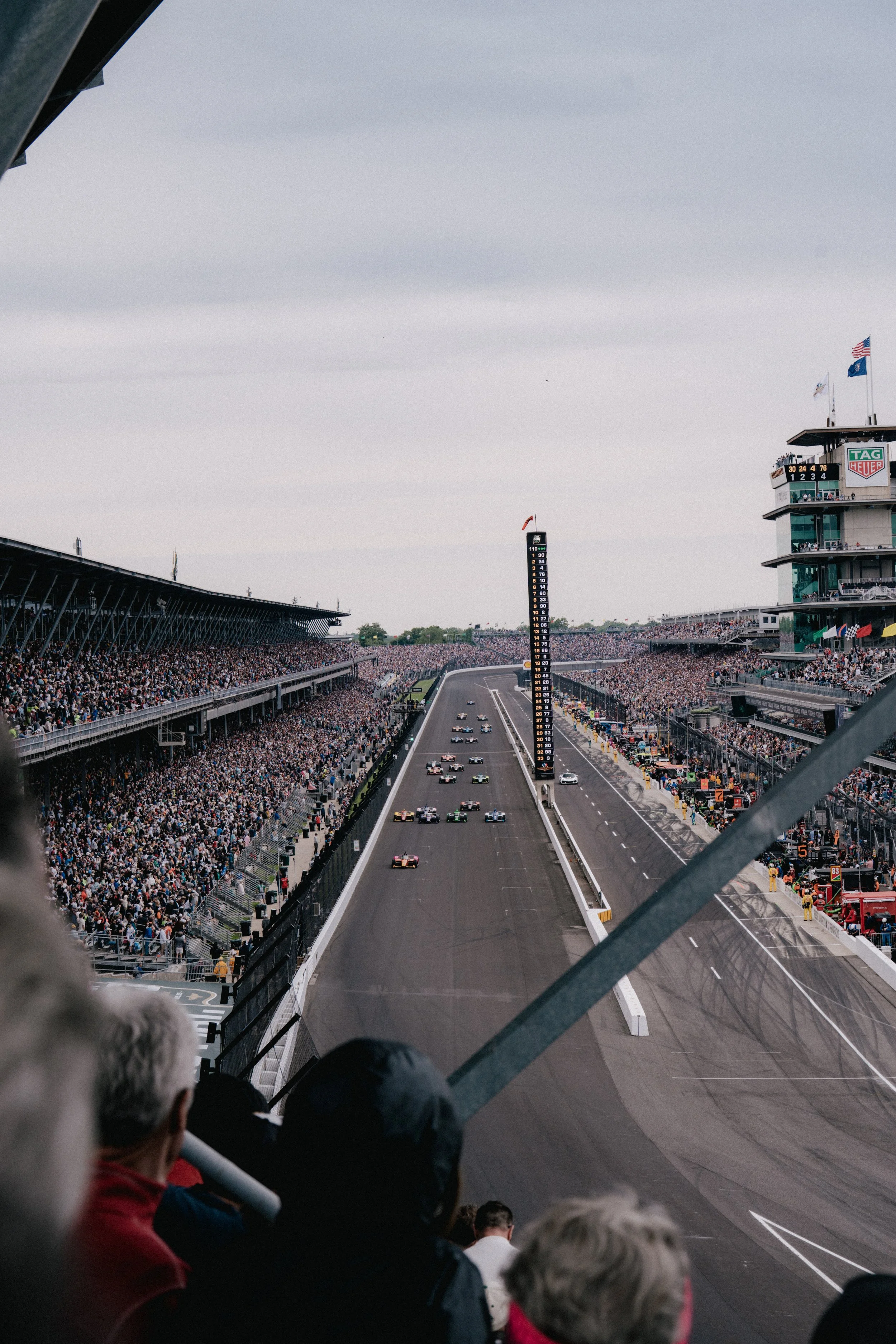 An outdoor car race at a large stadium, with multiple race cars on the track, a tall scoring tower displaying race positions, and a packed grandstand filled with spectators.