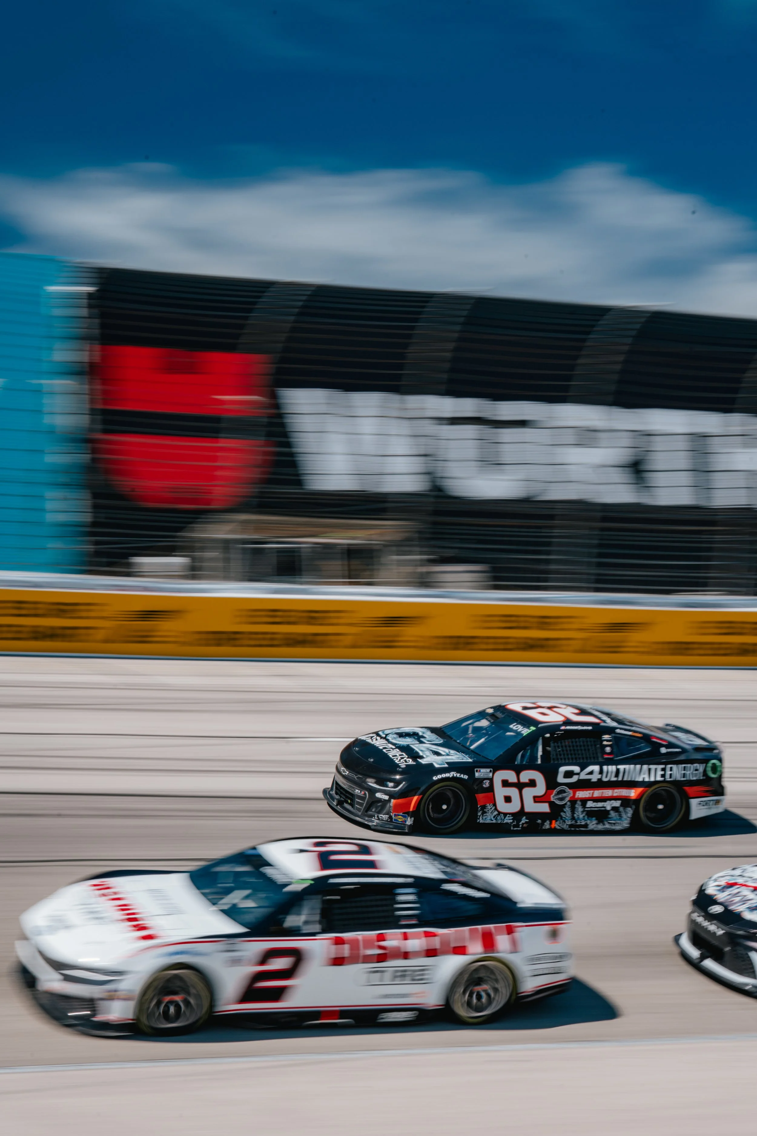 Two race cars on a track in front of a blurred background with a large sign that says 'MotorSpeedway.'
