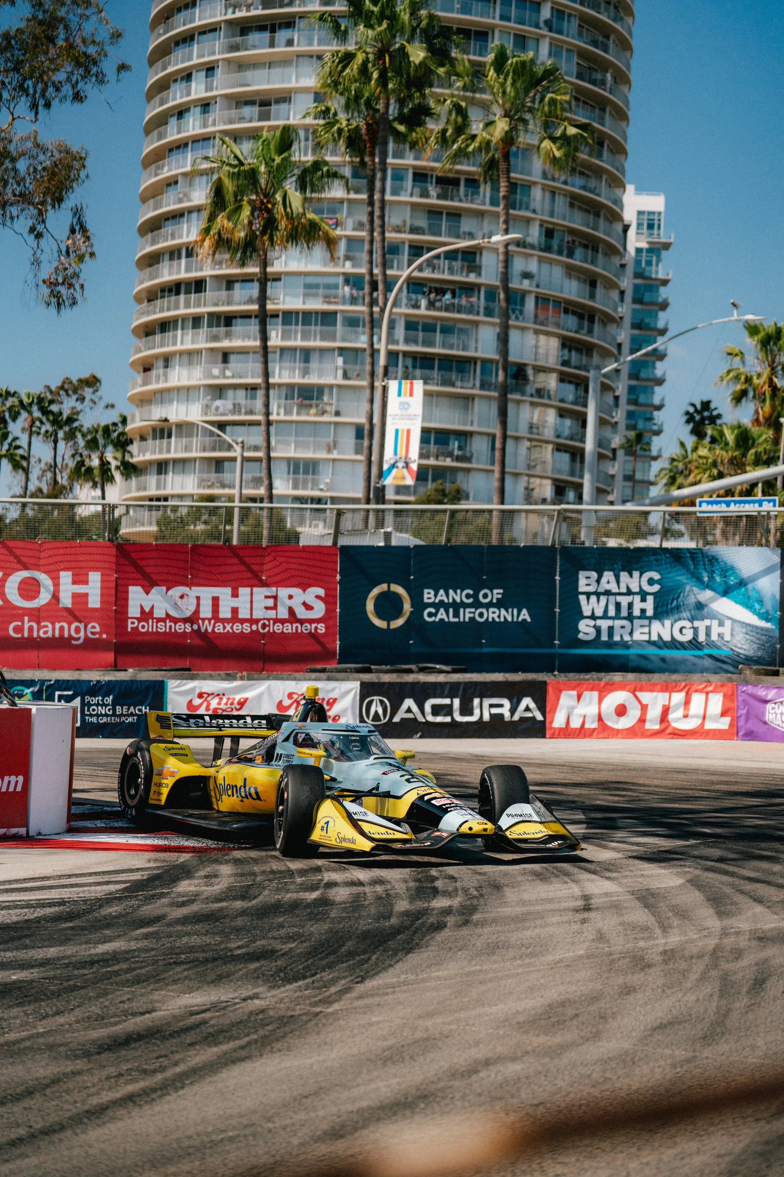 A yellow and black IndyCar racing on a street circuit with palm trees, tall buildings, and advertising banners in the background.