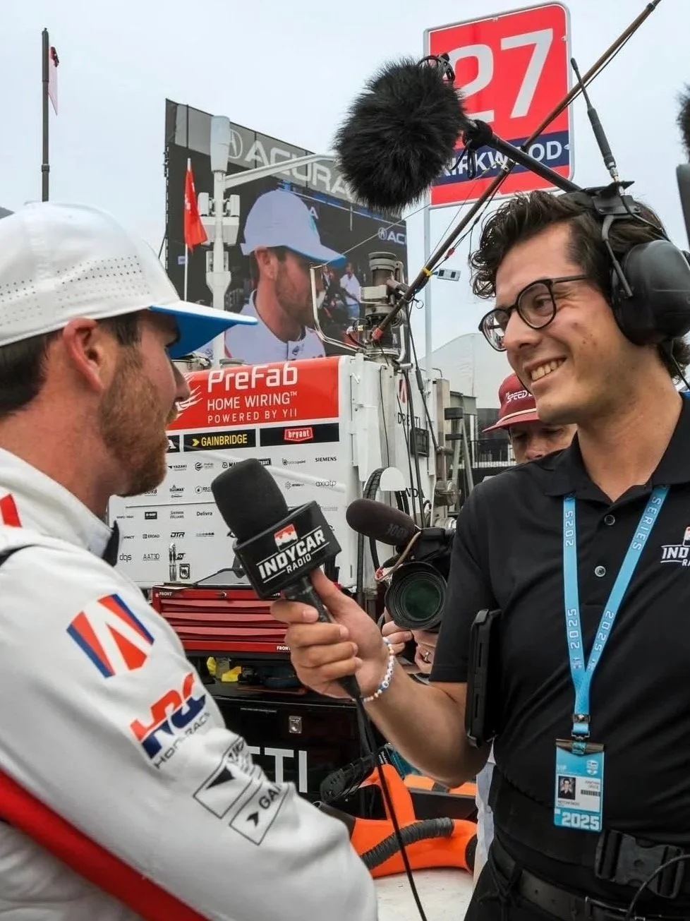 A race car driver being interviewed by a journalist with a large microphone and a camera around his neck at a racing event, with a large screen and team trucks in the background.