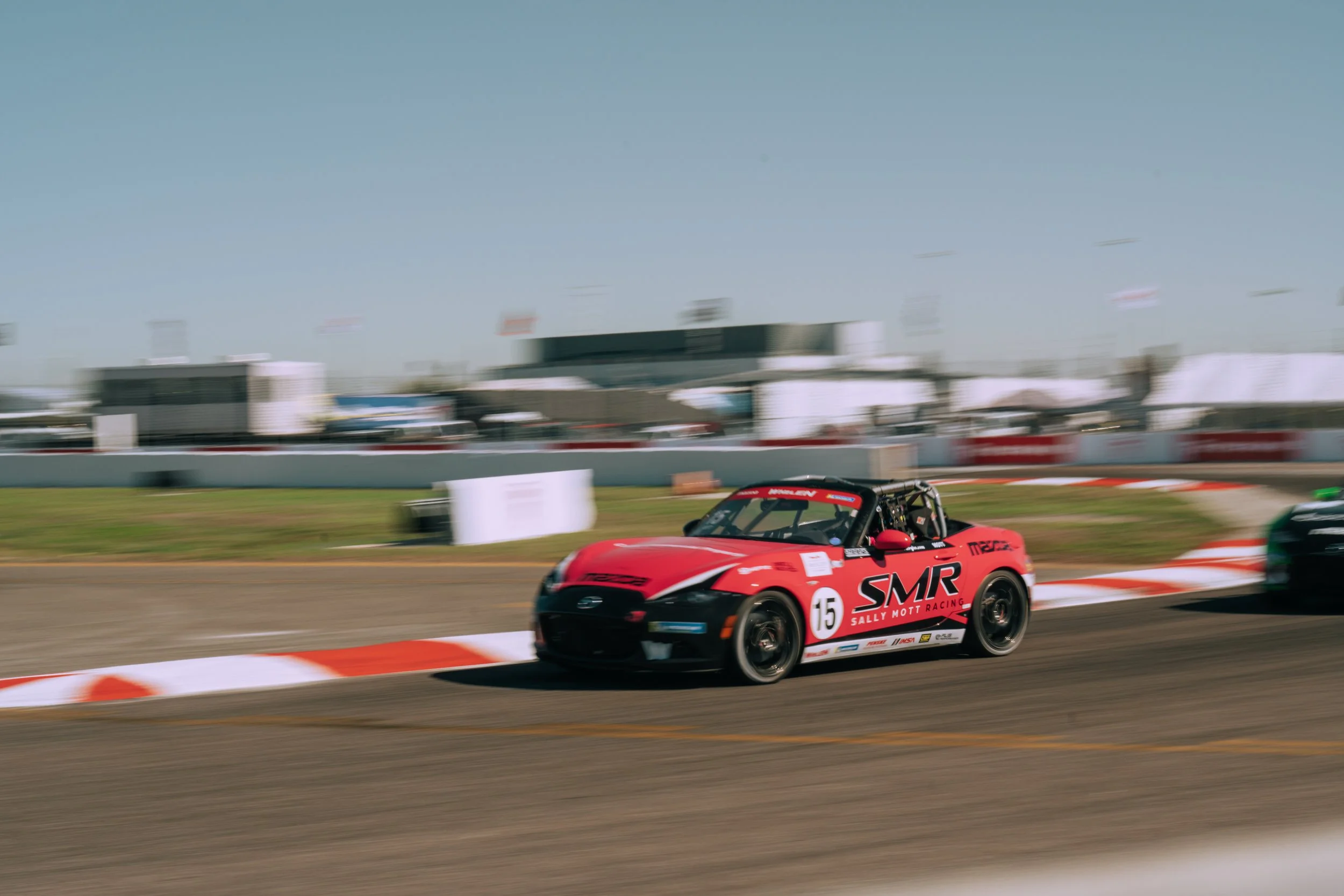 A red race car on a track with the number 15 and SMR logo, turning sharply with motion blur in the background.
