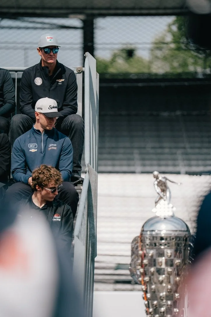 Three young men sitting on bleachers at a racing event, wearing team uniforms and caps, with a racing trophy visible in the foreground.