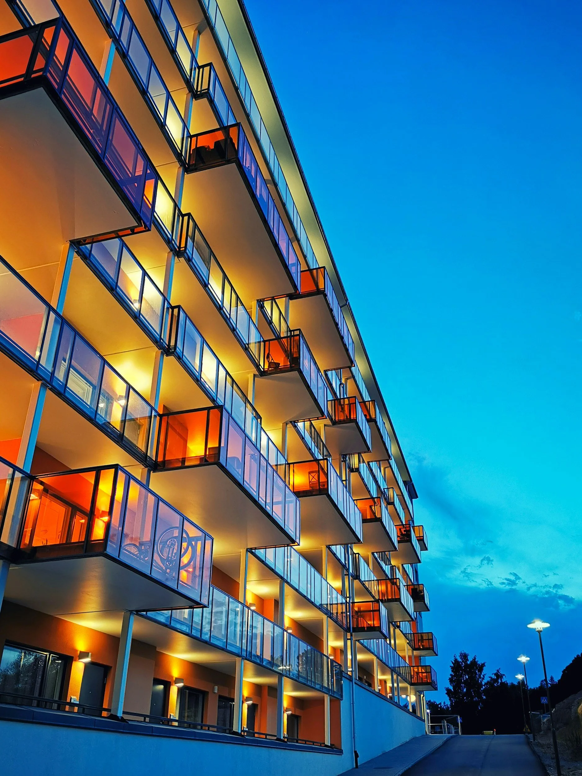 Modern multi-story apartment building with illuminated balconies at dusk, set against a blue sky with street lights.