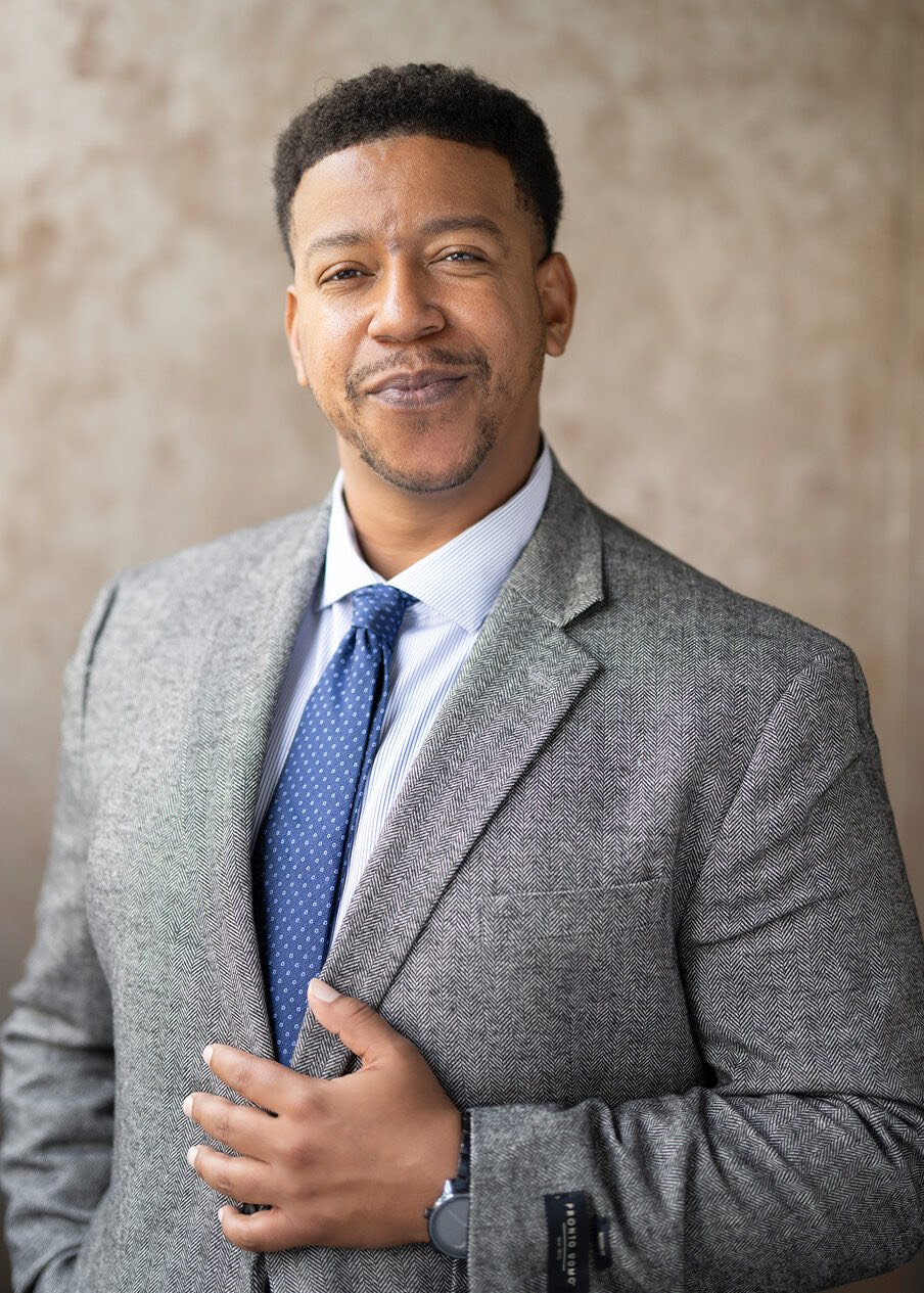 A confident man in a gray suit and blue tie standing against a neutral background.