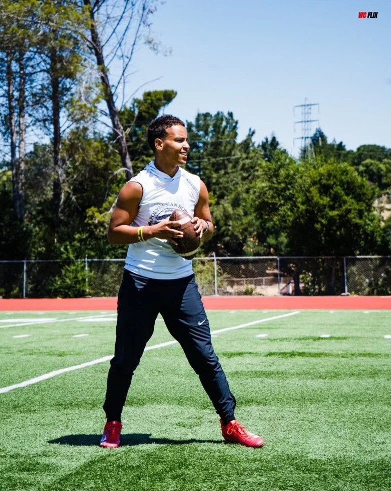 Young male athlete standing on a football field, holding a football, with trees and a clear blue sky in the background.