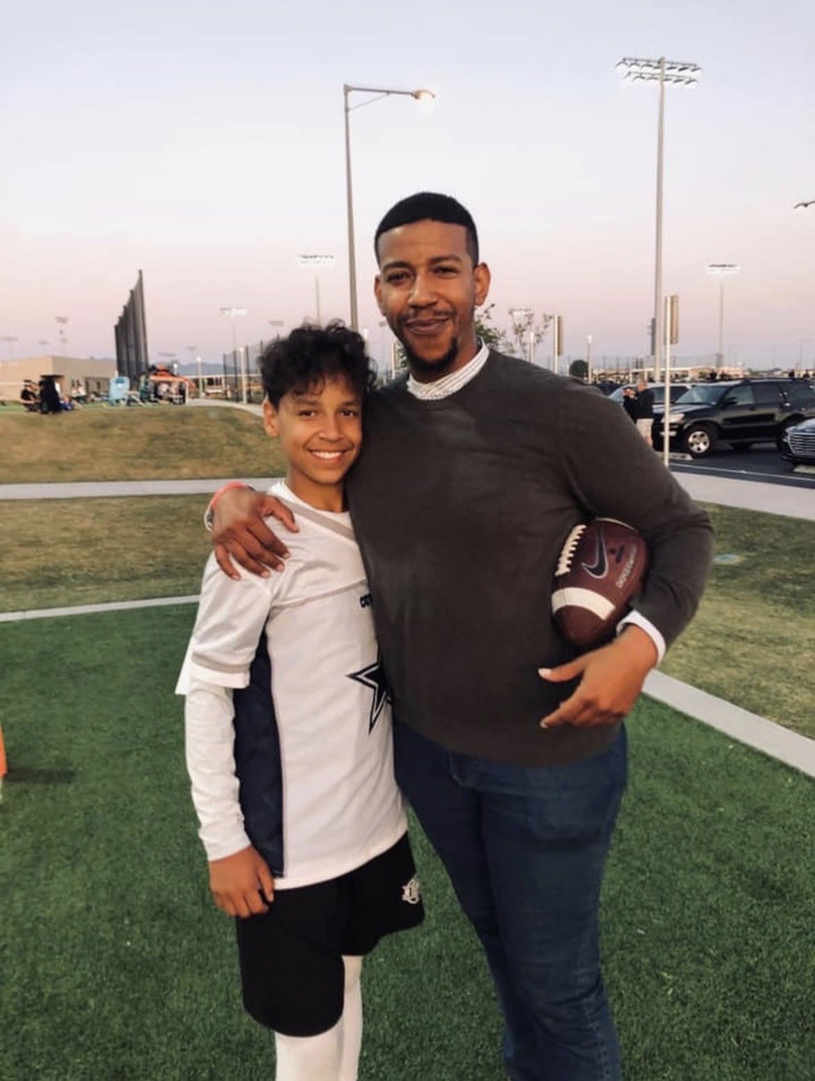 A Coach and his son standing on a sports field, smiling and posing for a photo, with the man holding a football under his arm.