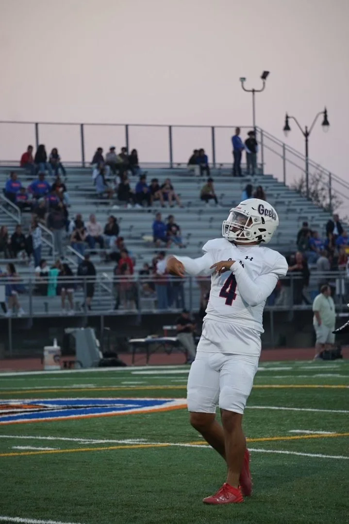 A football player in a white uniform and helmet with the word 'Gops' on it, standing on the field and looking to the side, with spectators in the background seated on bleachers and a few people standing near the fence.