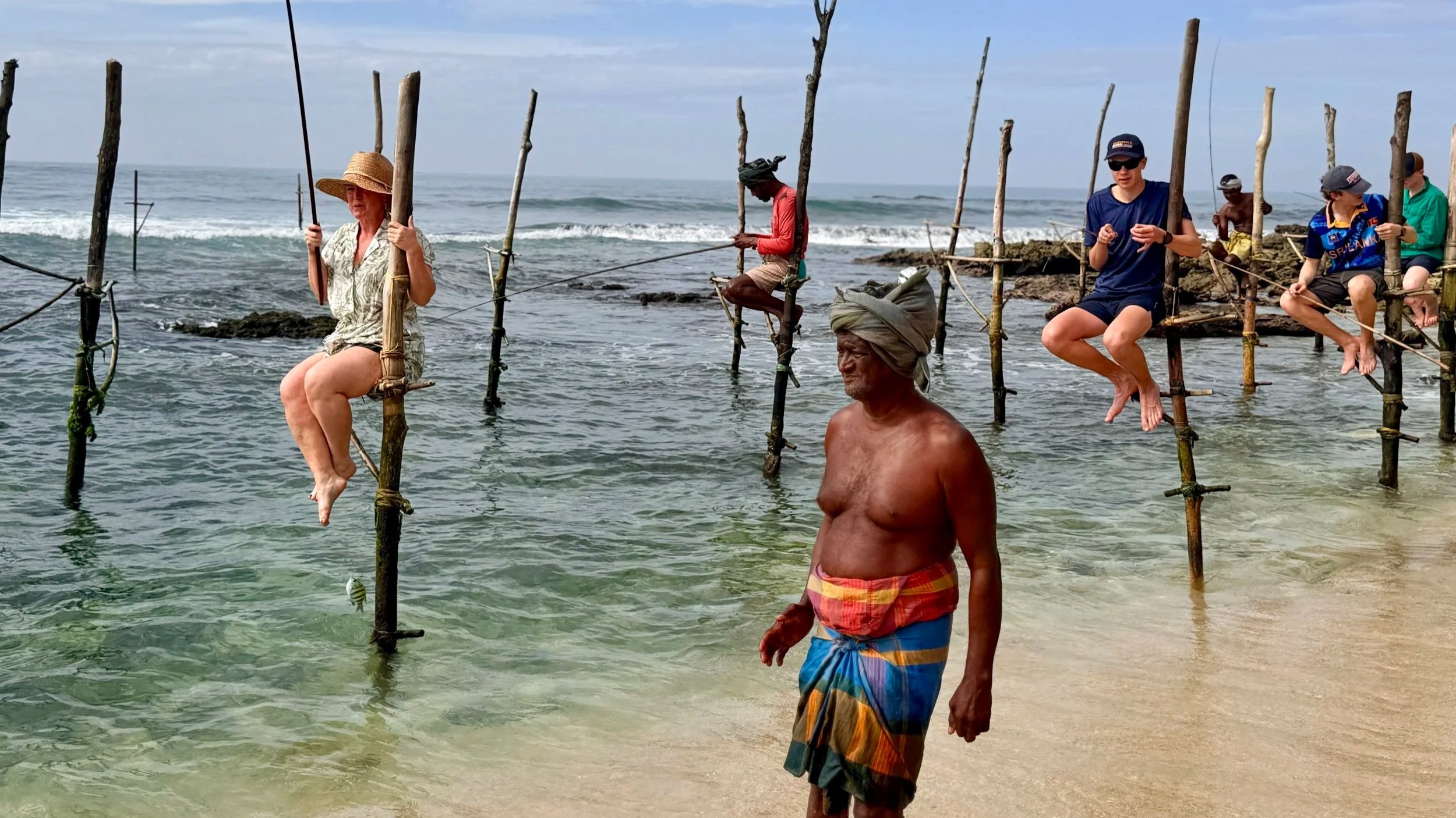 Group of people, including a woman with a straw hat sitting on a wooden structure and a man in colorful shorts, near a beach with water, rocks, and wooden poles, some people fishing.