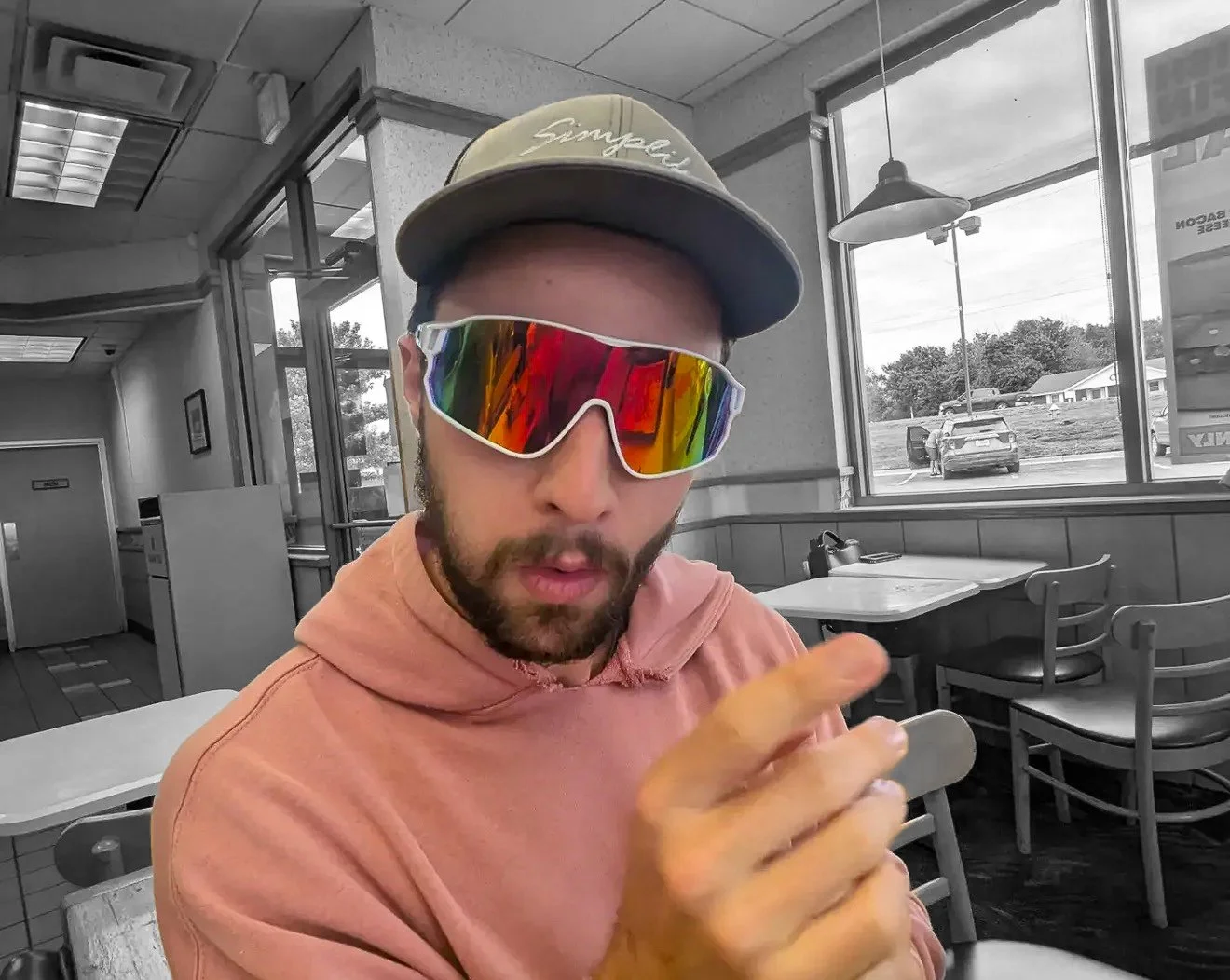 A man wearing a beige baseball cap and large futuristic sunglasses with rainbow reflective lenses inside a fast-food restaurant. He's wearing a pink hoodie and has a beard. The background shows a window, some tables, and chairs inside the restaurant, and parking lot outside.