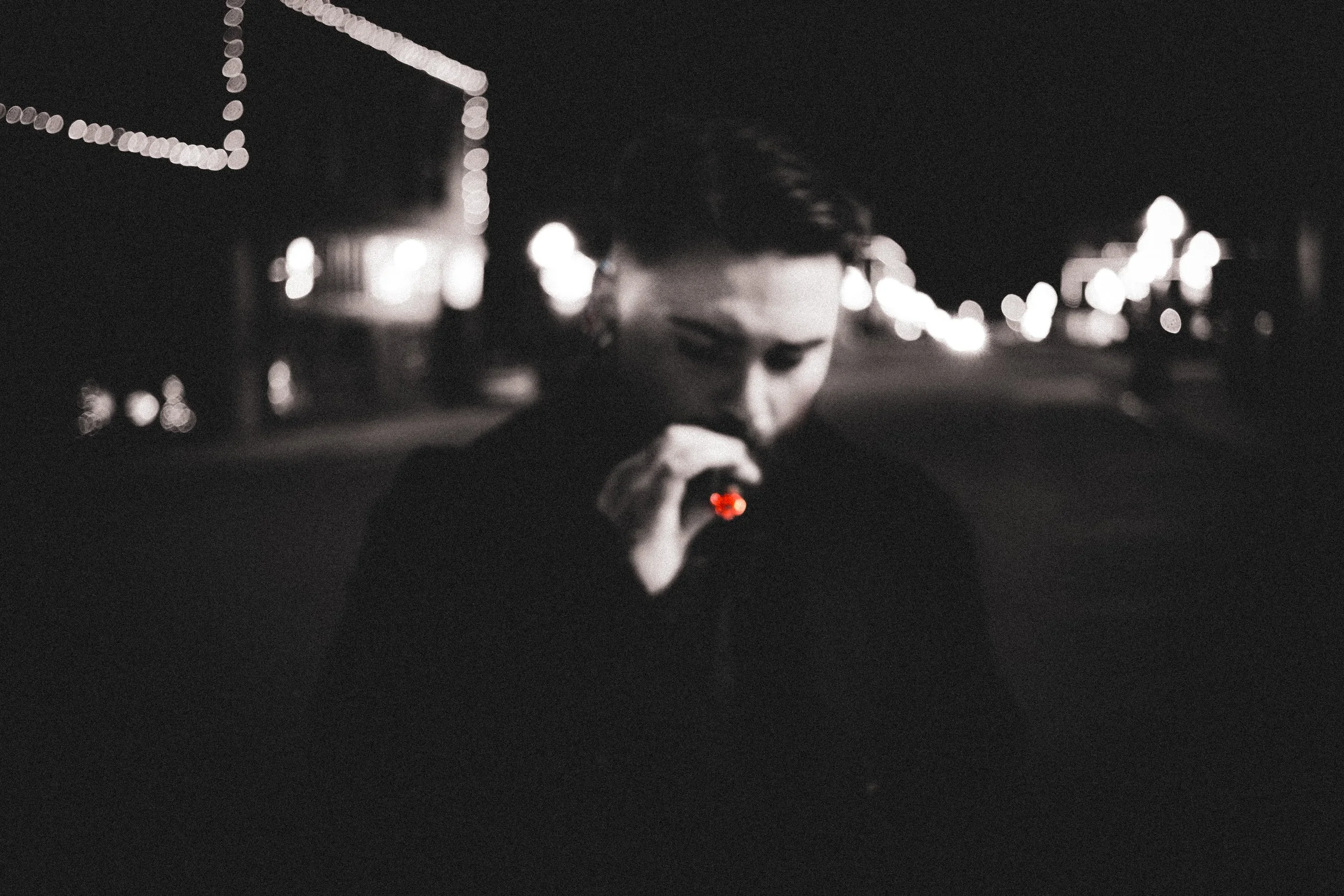 A person with glasses at night, holding a small red light or device in their hand, with blurred city lights in the background.