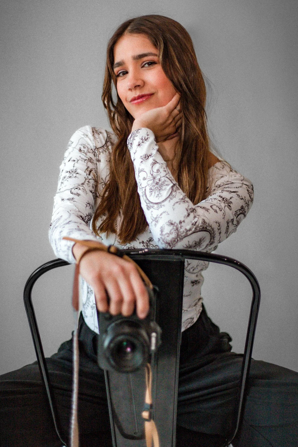A woman with long brown hair sitting on a black chair, smiling, with her chin resting on her hand. She is wearing a white blouse with floral patterns and black pants, and is holding a camera pointed at herself. The background is plain gray.