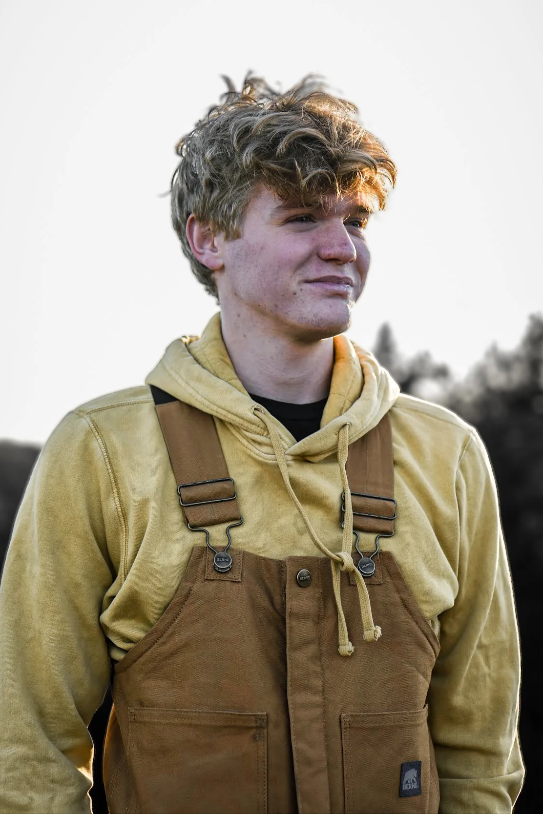 A young man with curly blonde hair stands outdoors, wearing a yellow hoodie and brown overall with straps.