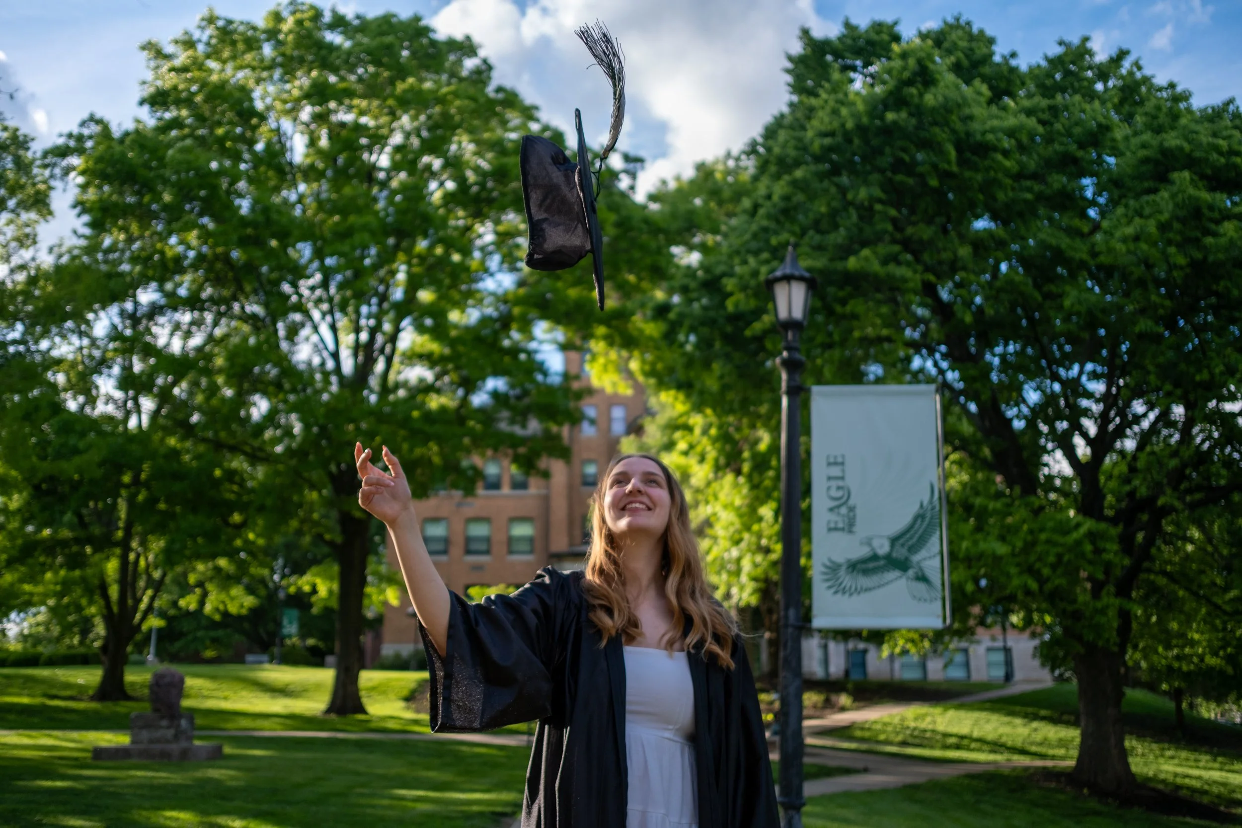 A young woman in academic regalia throwing a graduation cap in the air during a graduation ceremony outdoors in a park with green trees and a lamp post.