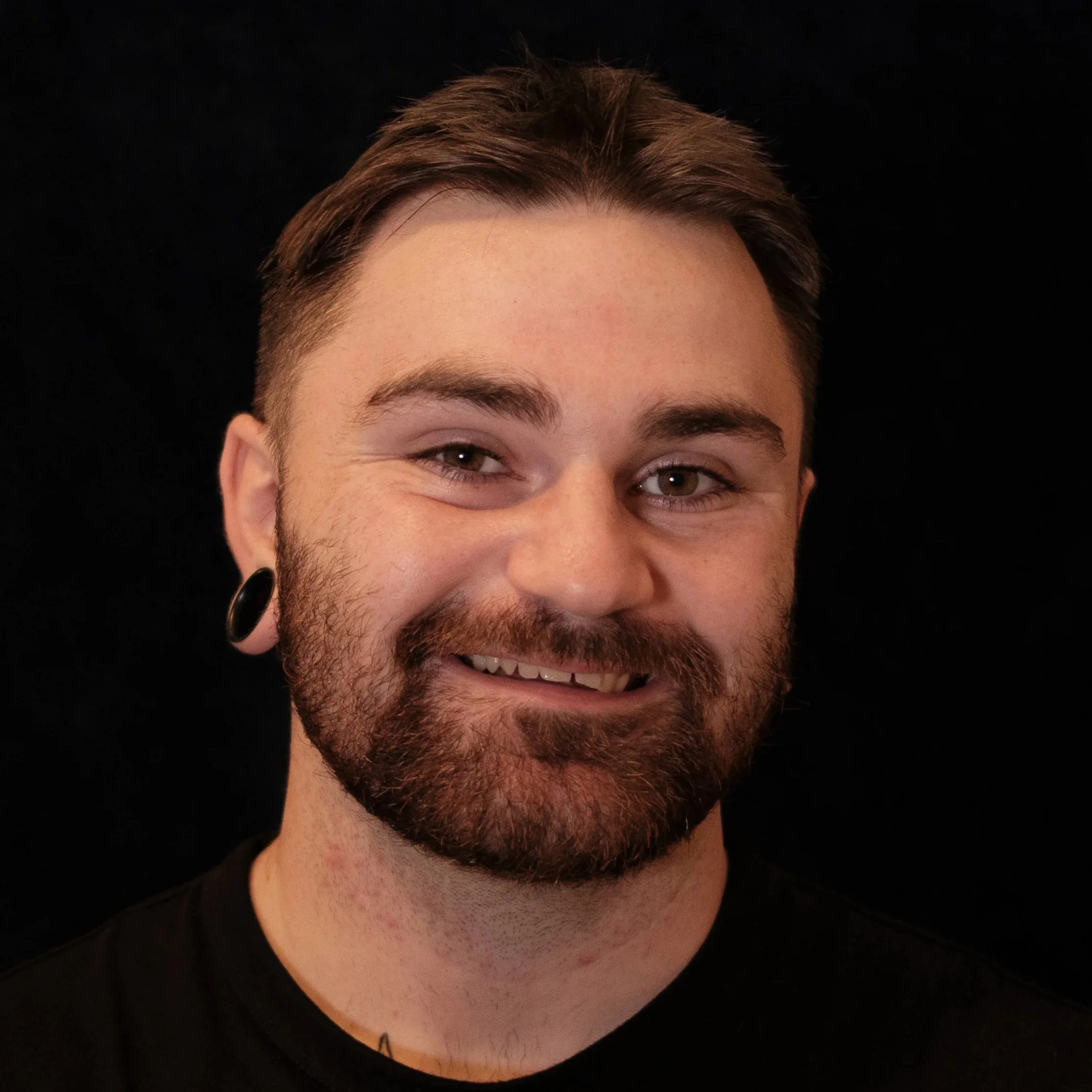 Close-up of a smiling young man with short brown hair, beard, and a black earring, against a black background.