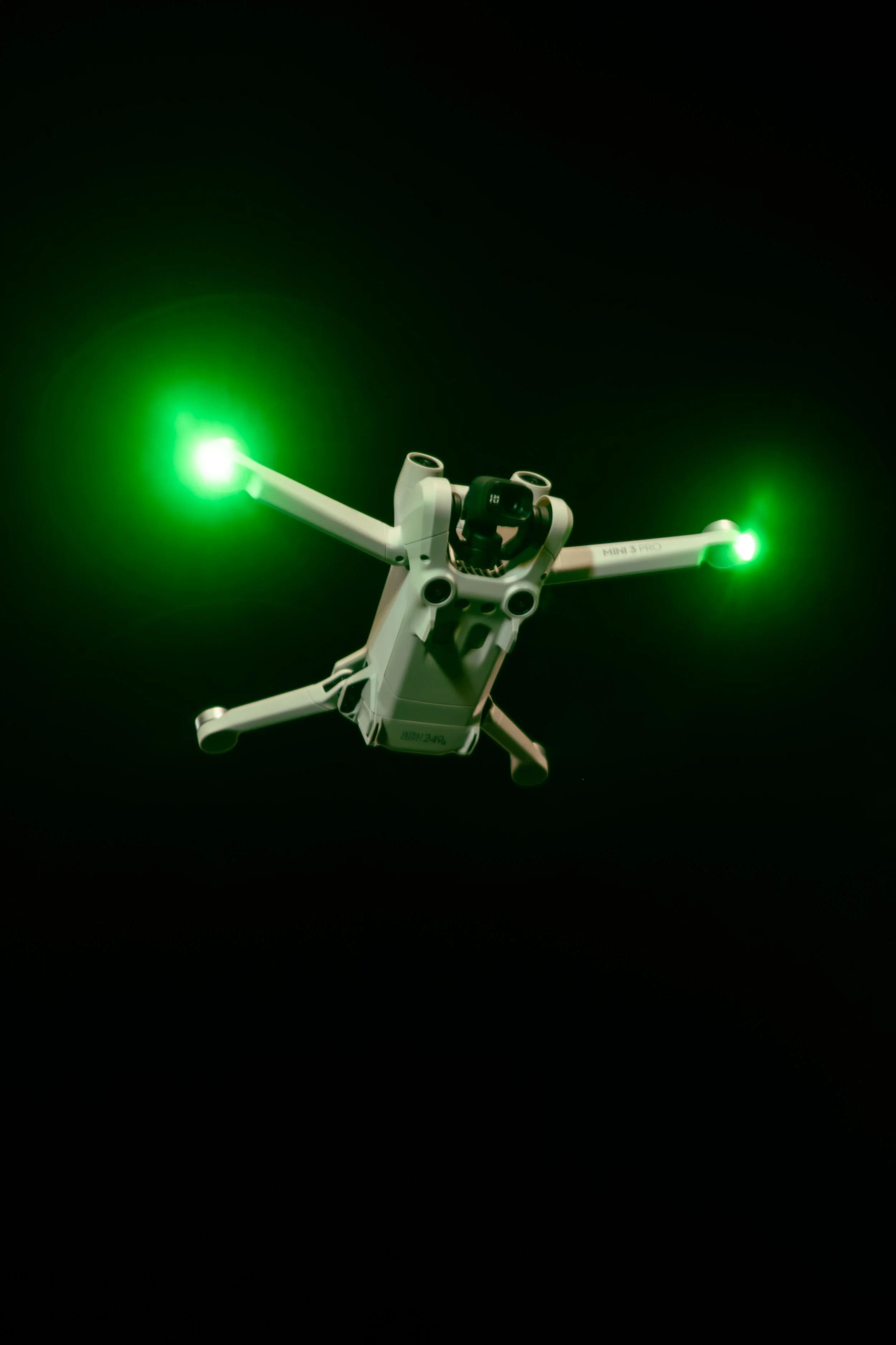 A white drone with green lights on its arms flying in a dark environment.