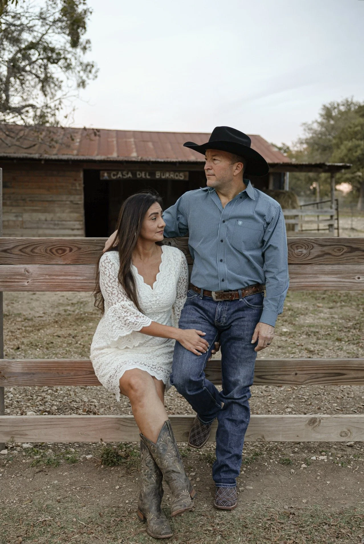 A man and a woman sit on a wooden fence in a rural area with a barn in the background. The man, wearing a cowboy hat and western attire, looks at the woman. The woman, dressed in a white lace dress and cowgirl boots, looks at the man, creating a romantic scene.