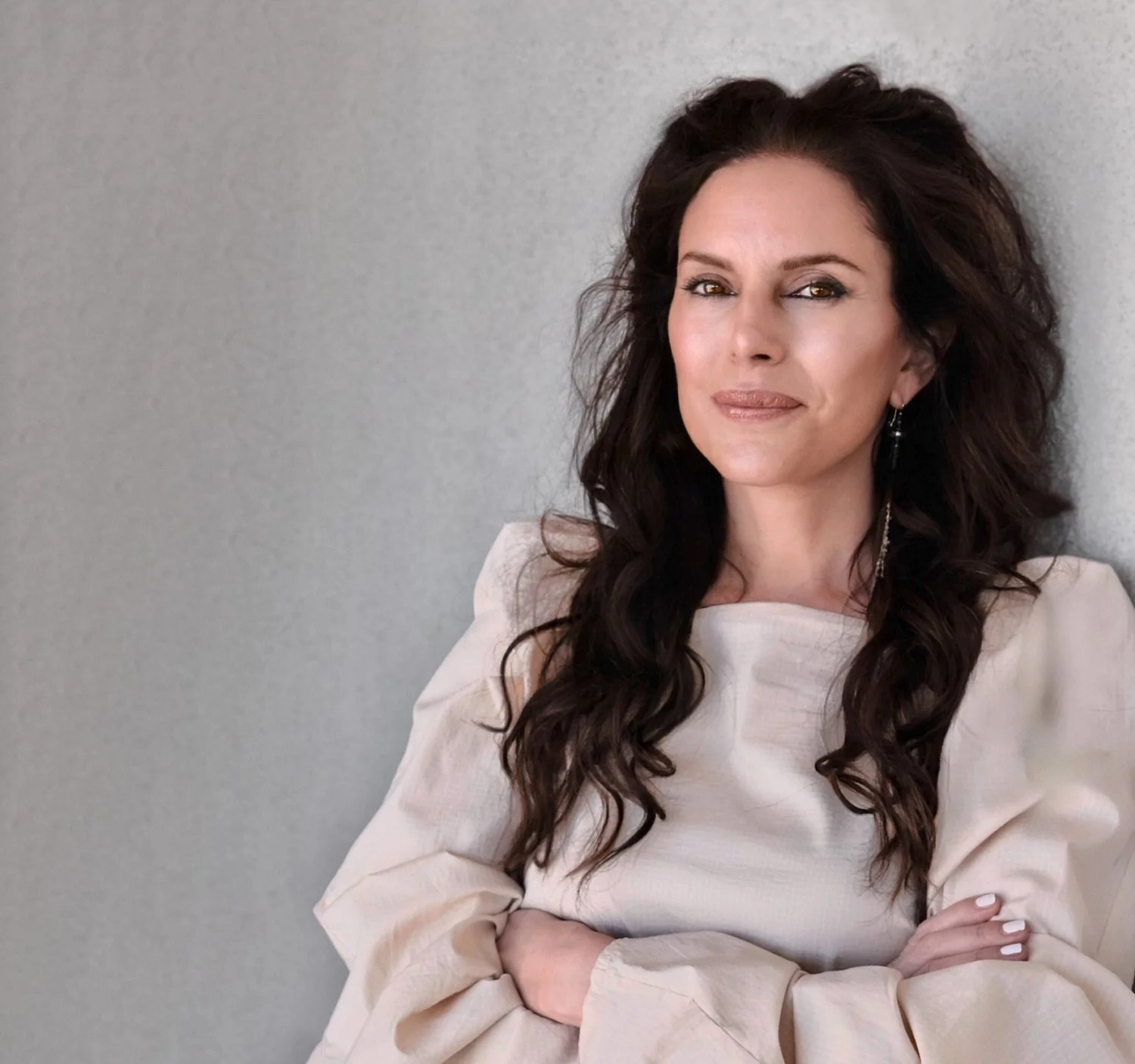 A woman with long dark wavy hair and earrings, standing against a light gray wall, wearing a light beige or off-white top, with her arms crossed.
