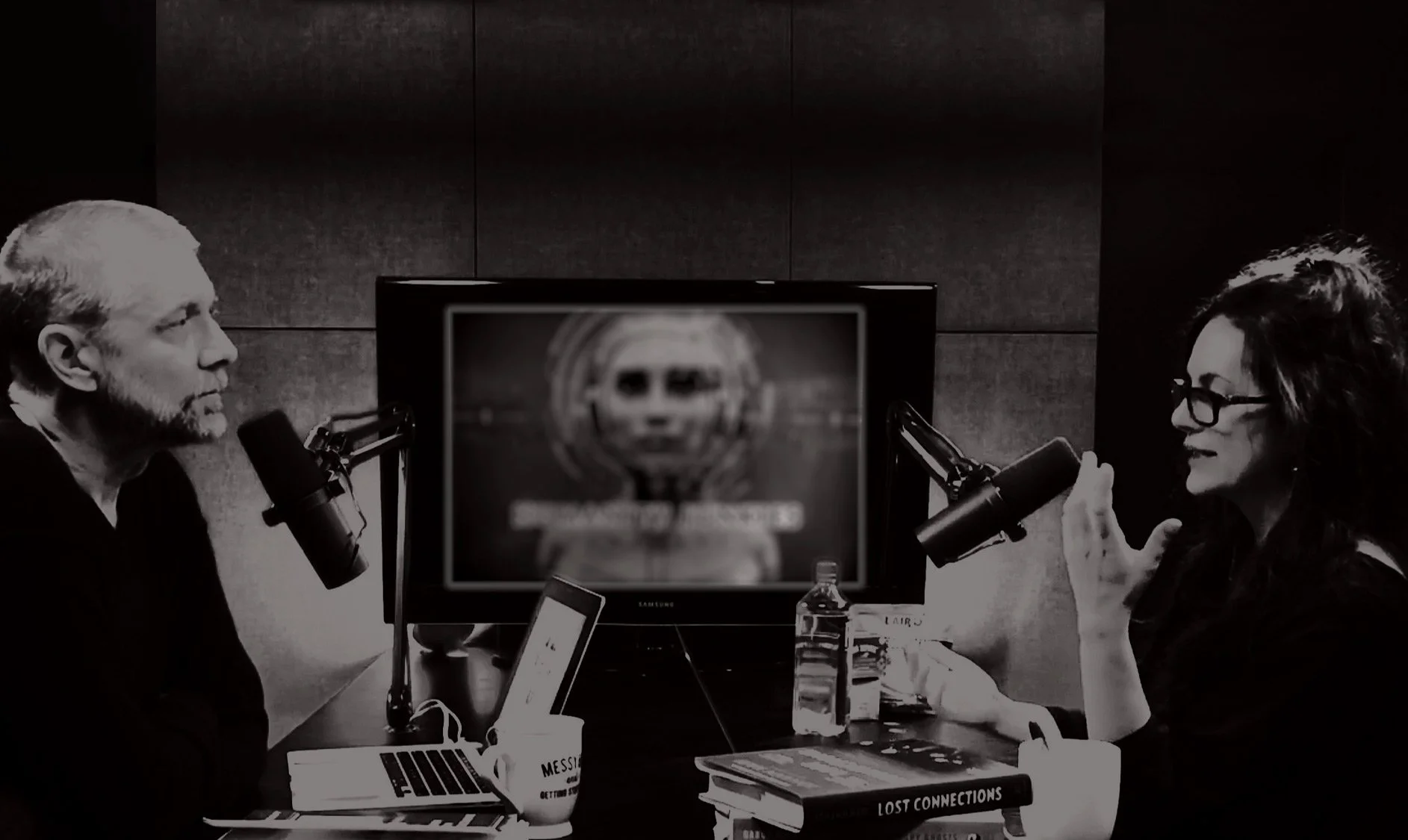 Two people speaking into microphones in a podcast studio, with a blurred Chappie AI logo on a monitor in the background and books on the table.