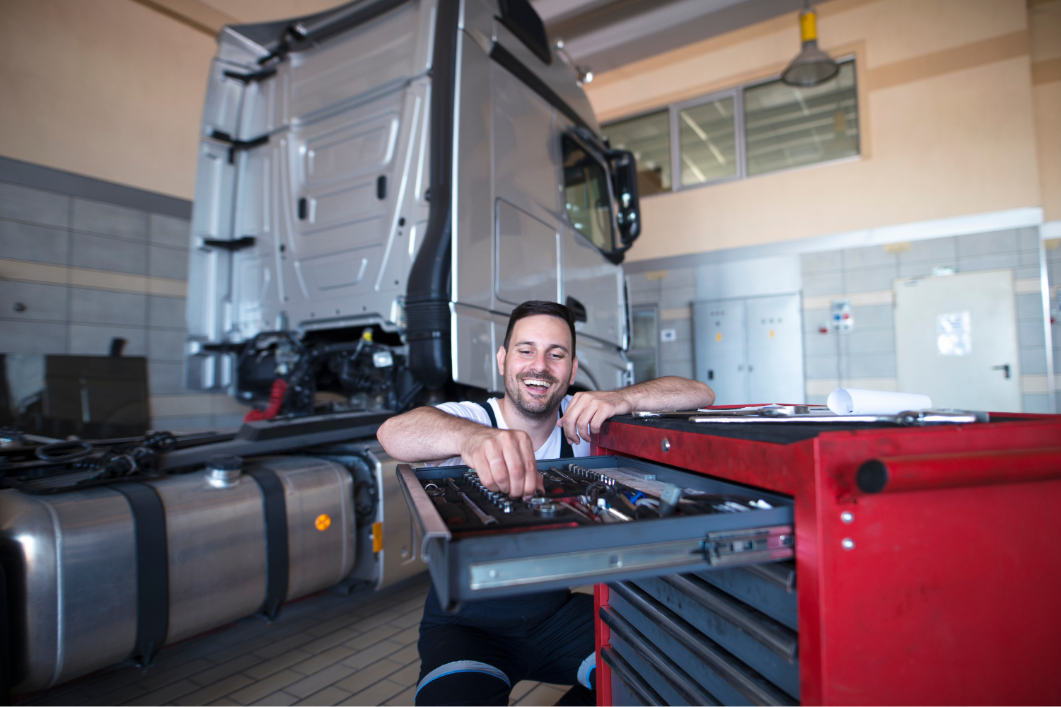 A man working on a tool chest inside a garage or workshop, with a large semi-truck behind him, smiling and looking at the tools.