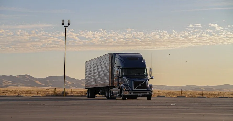 A large black semi-truck driving on a highway in a desert landscape with mountains in the background under a partly cloudy sky.